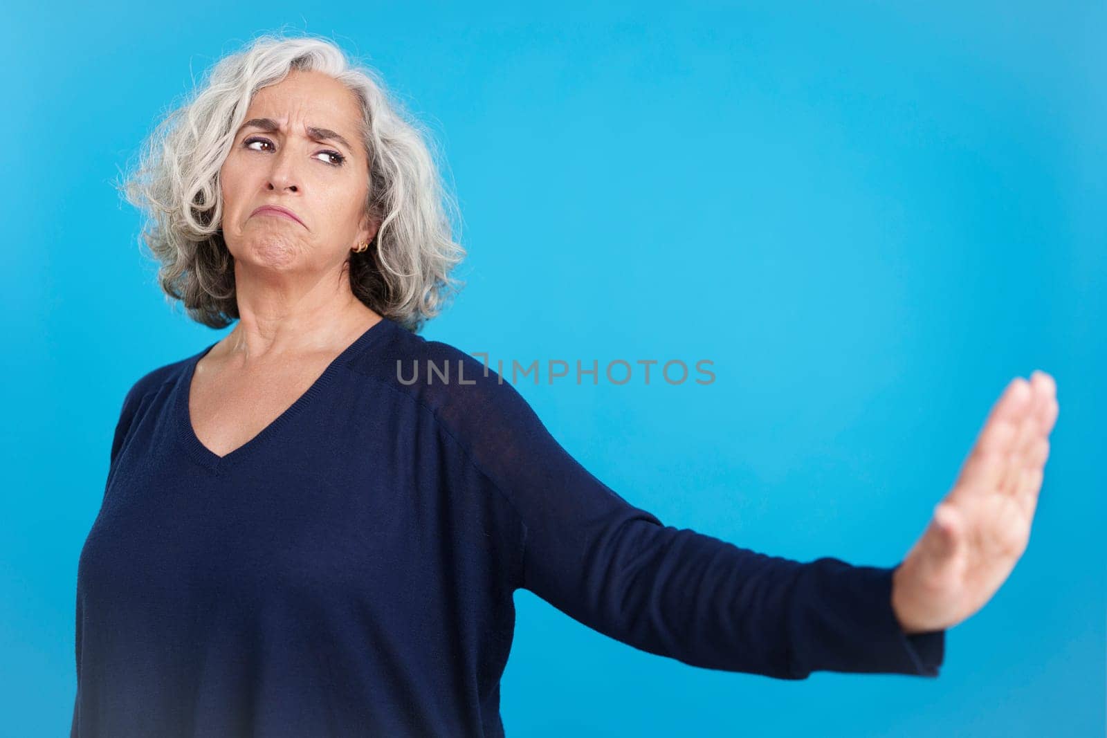 Studio portrait with blue background of a mature woman gesturing that she wants to know nothing by pushing away with her hand