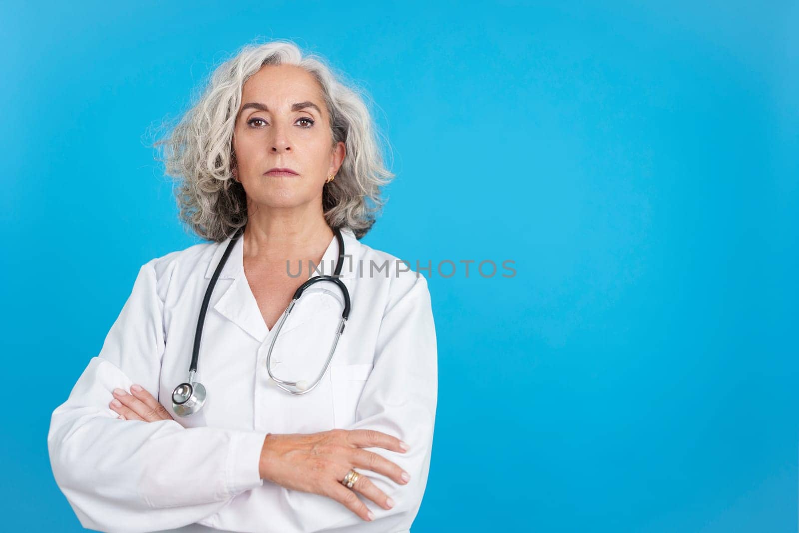 Studio portrait with blue background of a mature female doctor in white lab coat with crossed arms looking seriously at camera