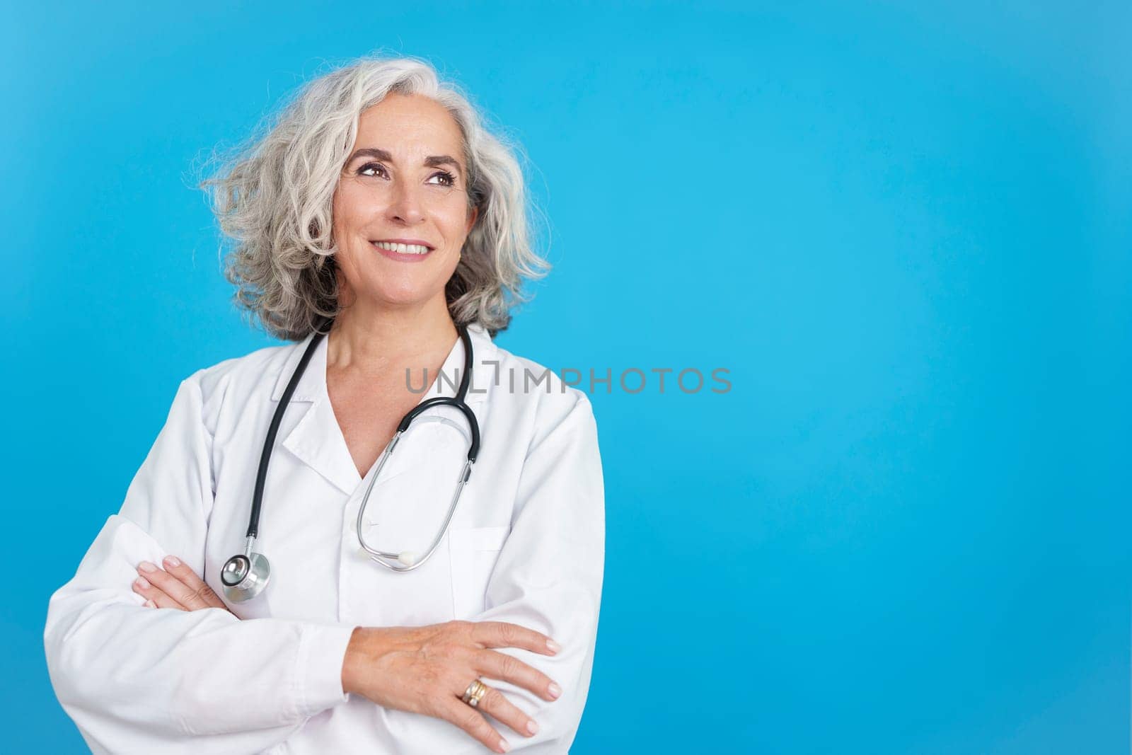 Studio portrait with blue background of a Mature female doctor in white coat looking up with the arms crossed