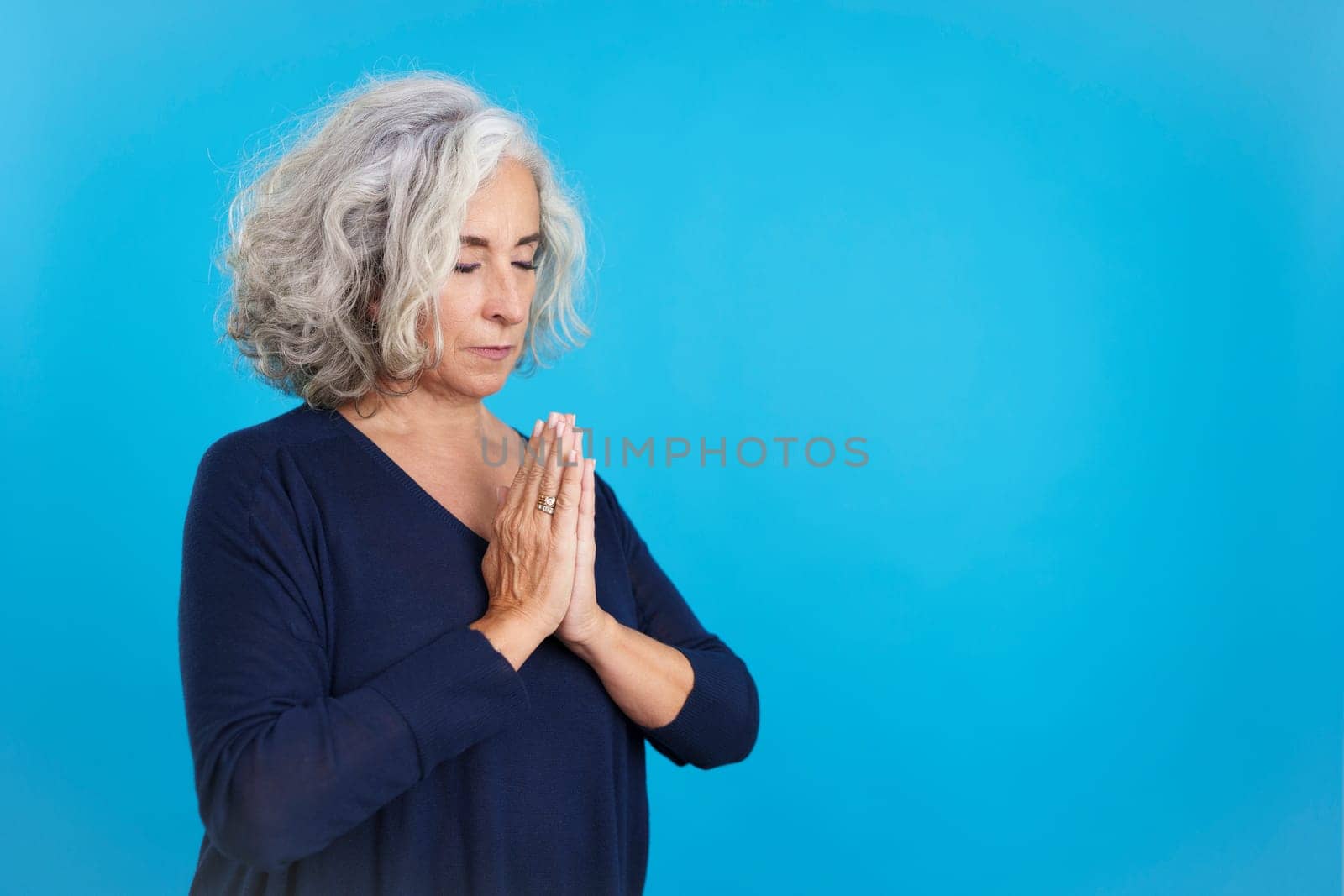 Studio portrait with blue background of a mature woman praying with folded hands and closed eyes