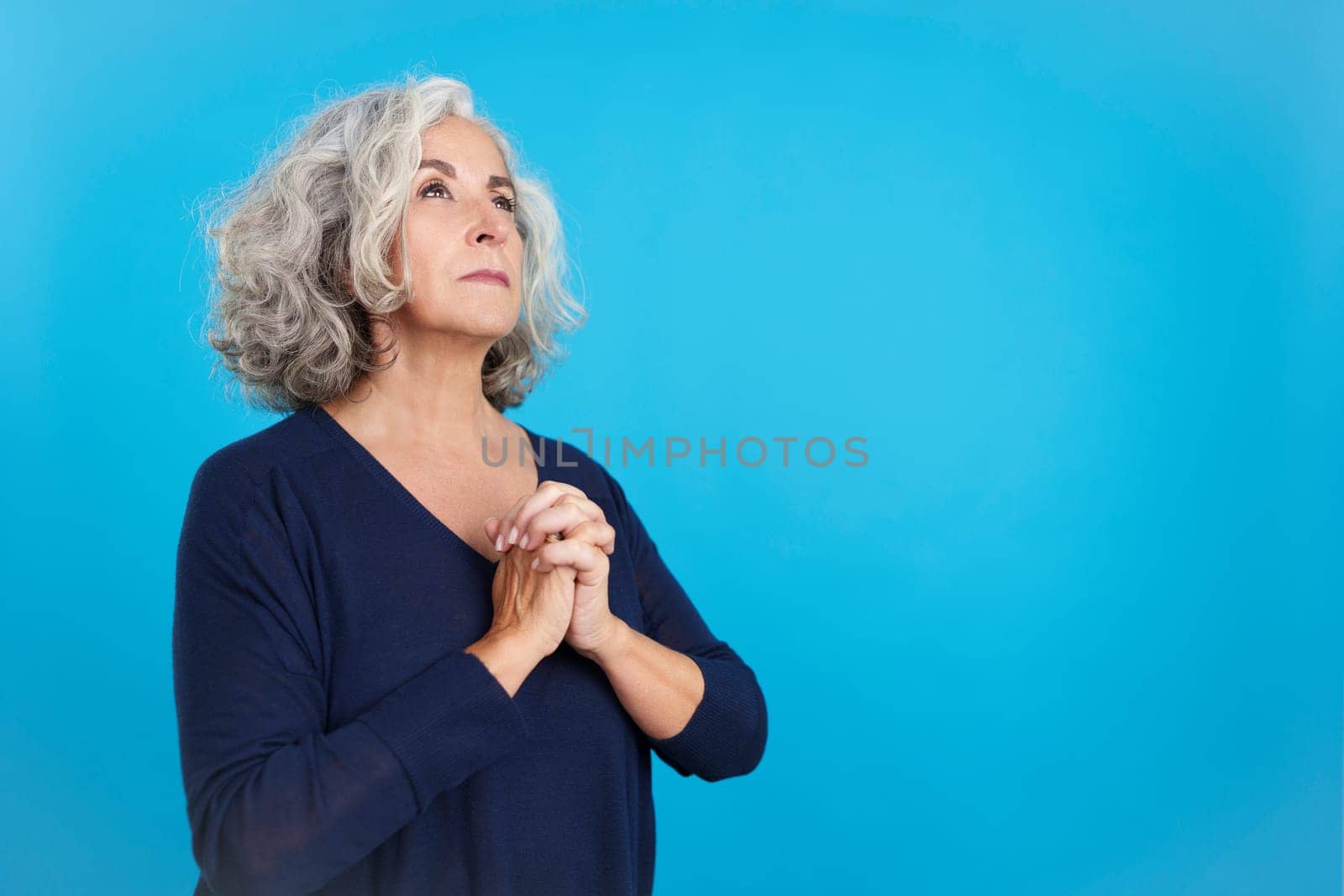 Studio portrait with blue background of a mature woman looking up while praying with folded hands