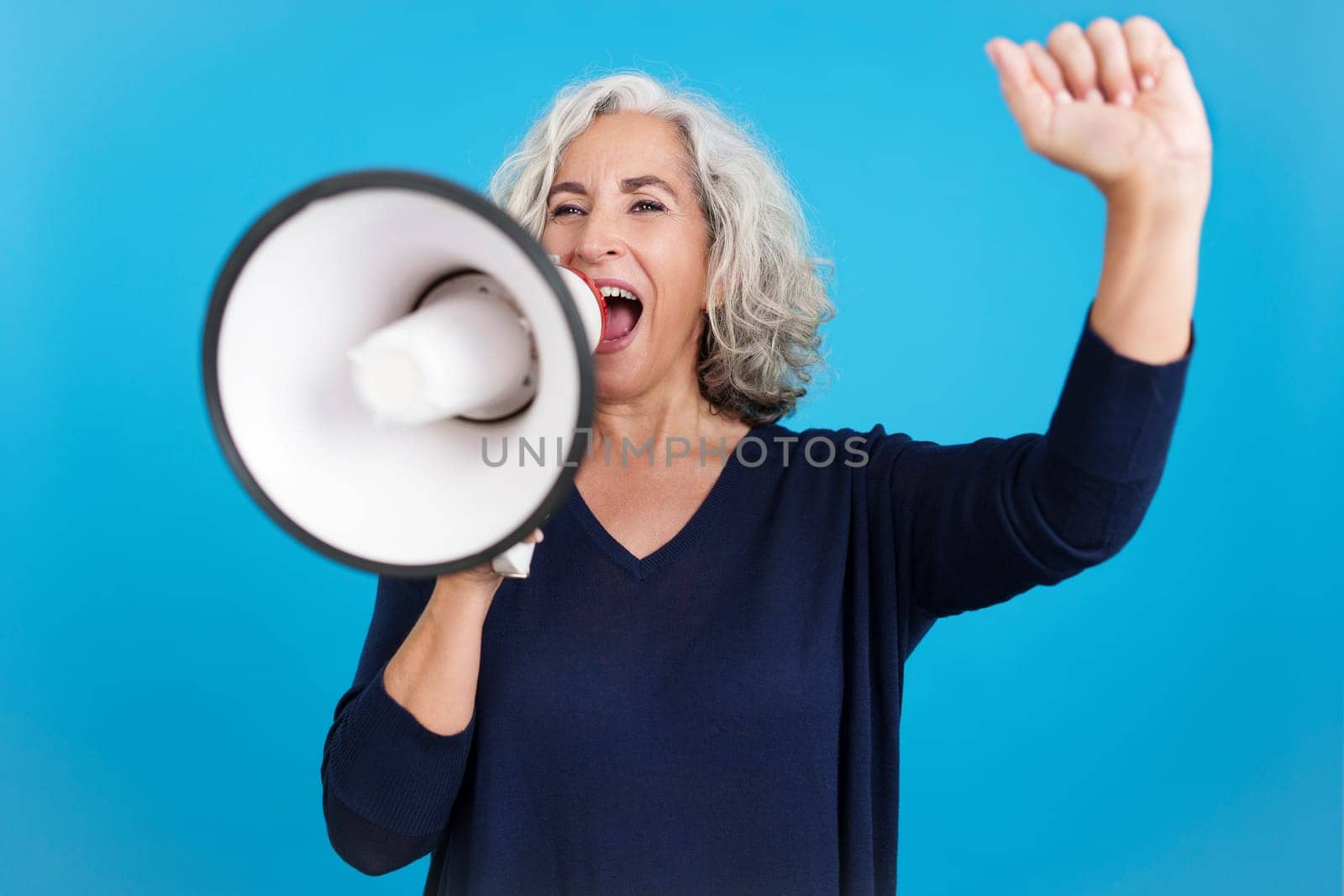 Studio portrait with blue background of a mature woman raising the fist and protesting with a loudspeaker