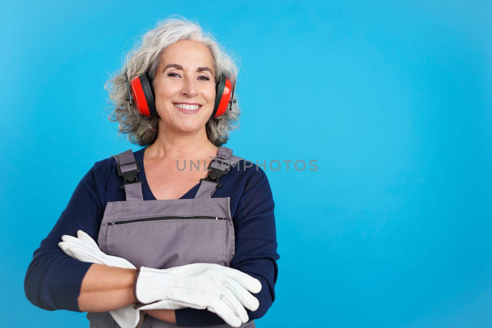 Studio portrait with blue background of an aged female carpentry worker with tools and work uniform smiling at the camera with arms crossed