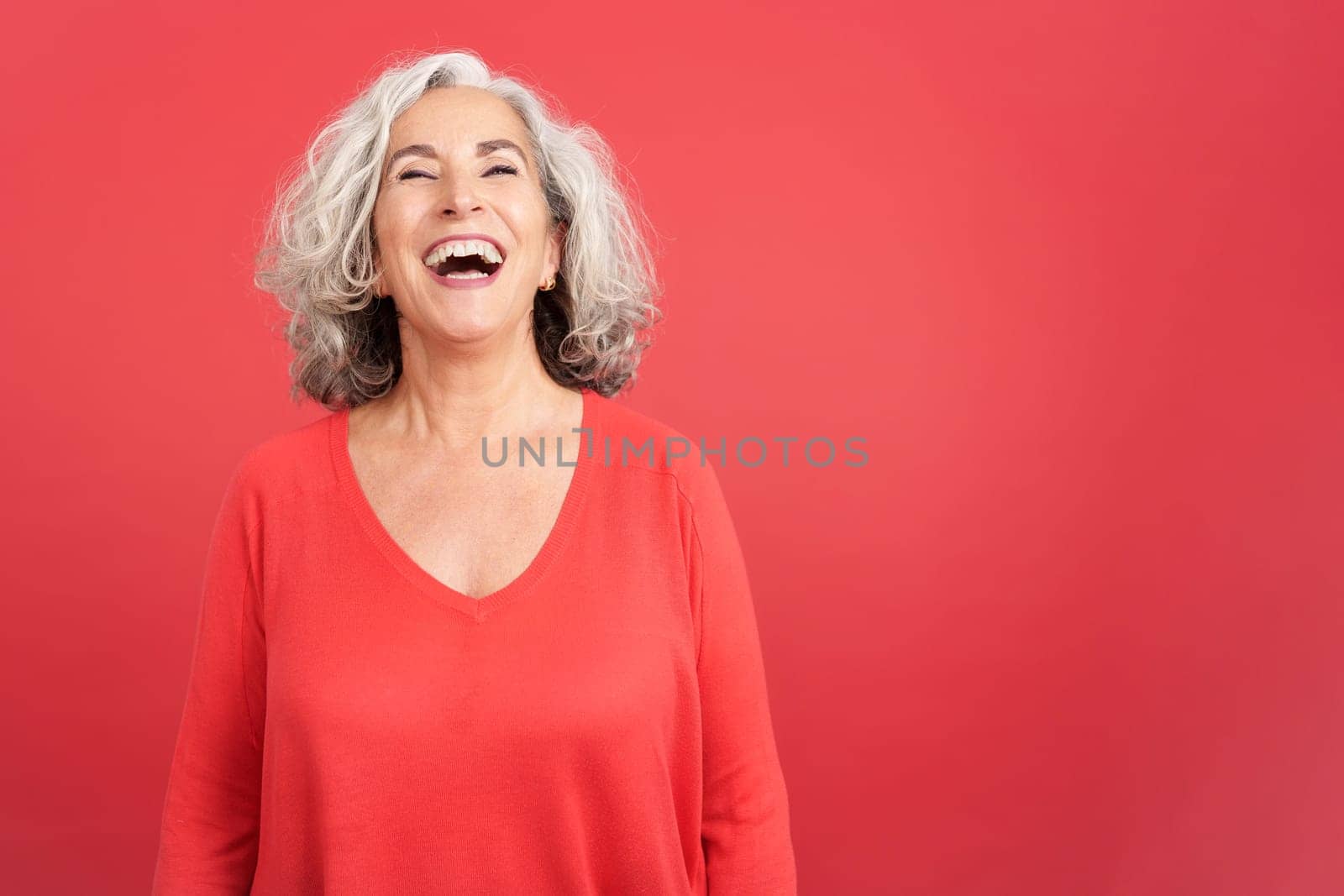 Studio portrait with red background of a mature woman in red shirt laughing while standing