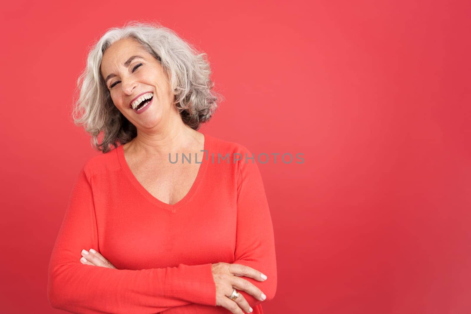 Studio portrait with red background of a happy aged woman standing with arms crossed