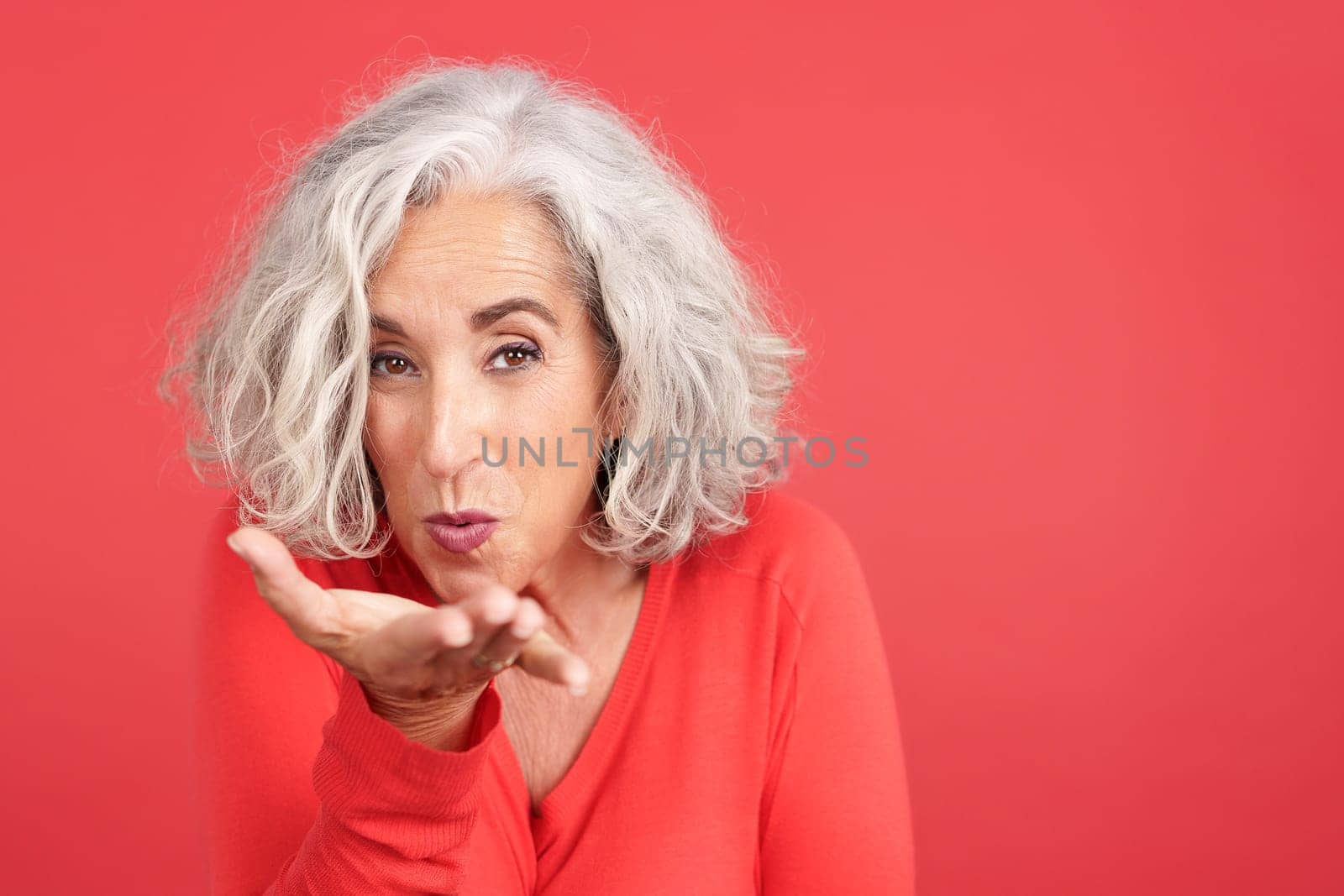 Studio portrait with close up view and red background of a mature woman blowing a kiss to the camera