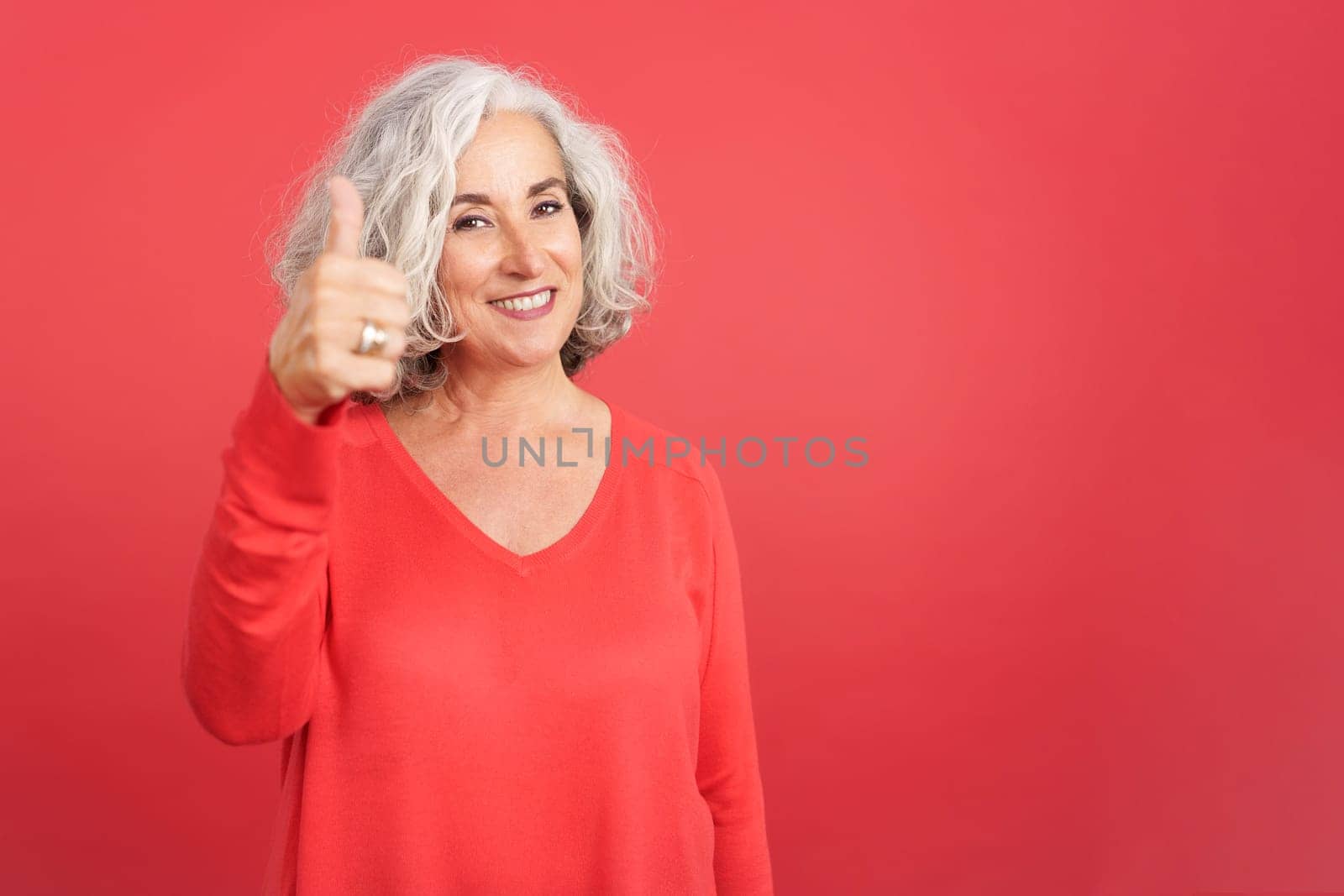 Studio portrait with red background of a mature woman gesturing agreement raising the thumb