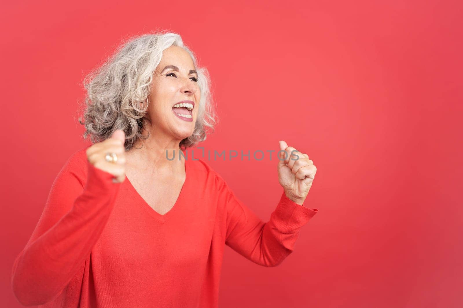 Studio portrait with red background of an excited mature woman celebrating while raising the fists