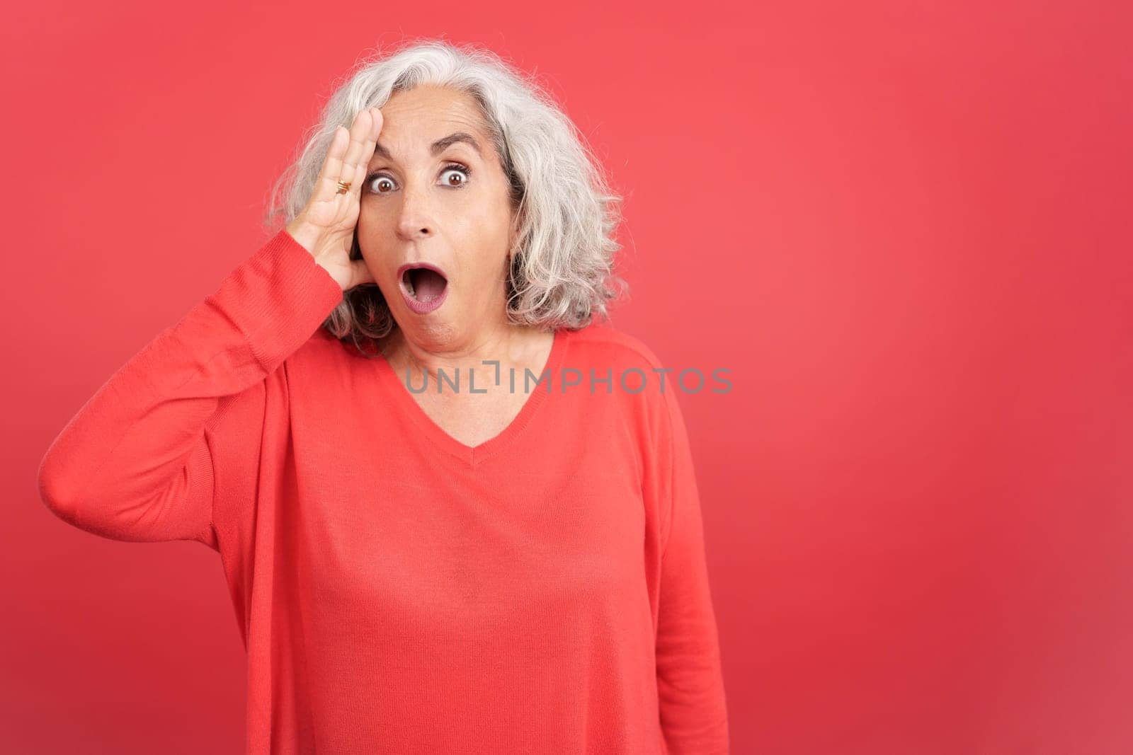 Studio portrait with red background of a mature woman looking at camera with a surprised look