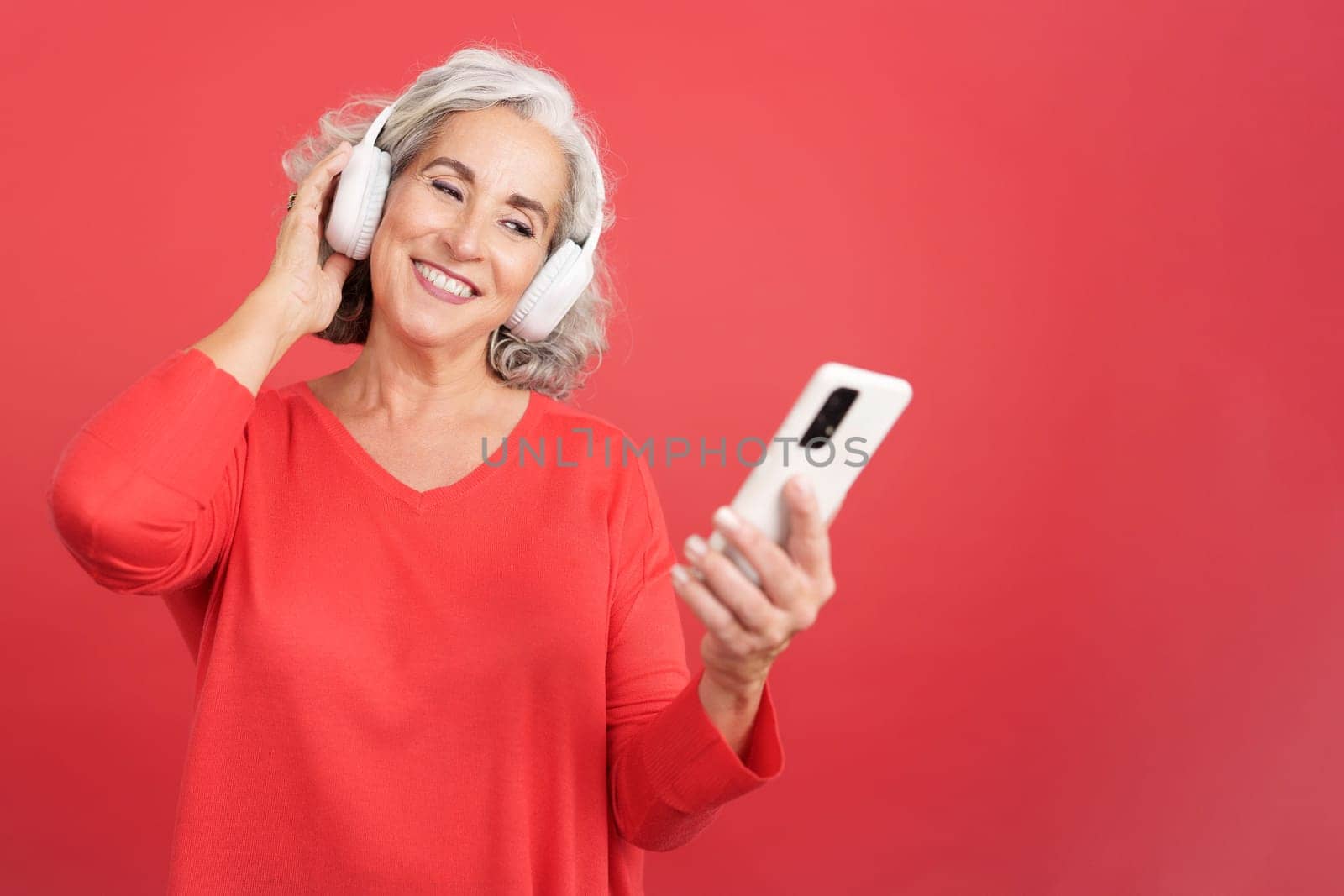 Studio portrait with red background of a mature woman listening to music with a mobile and headphones