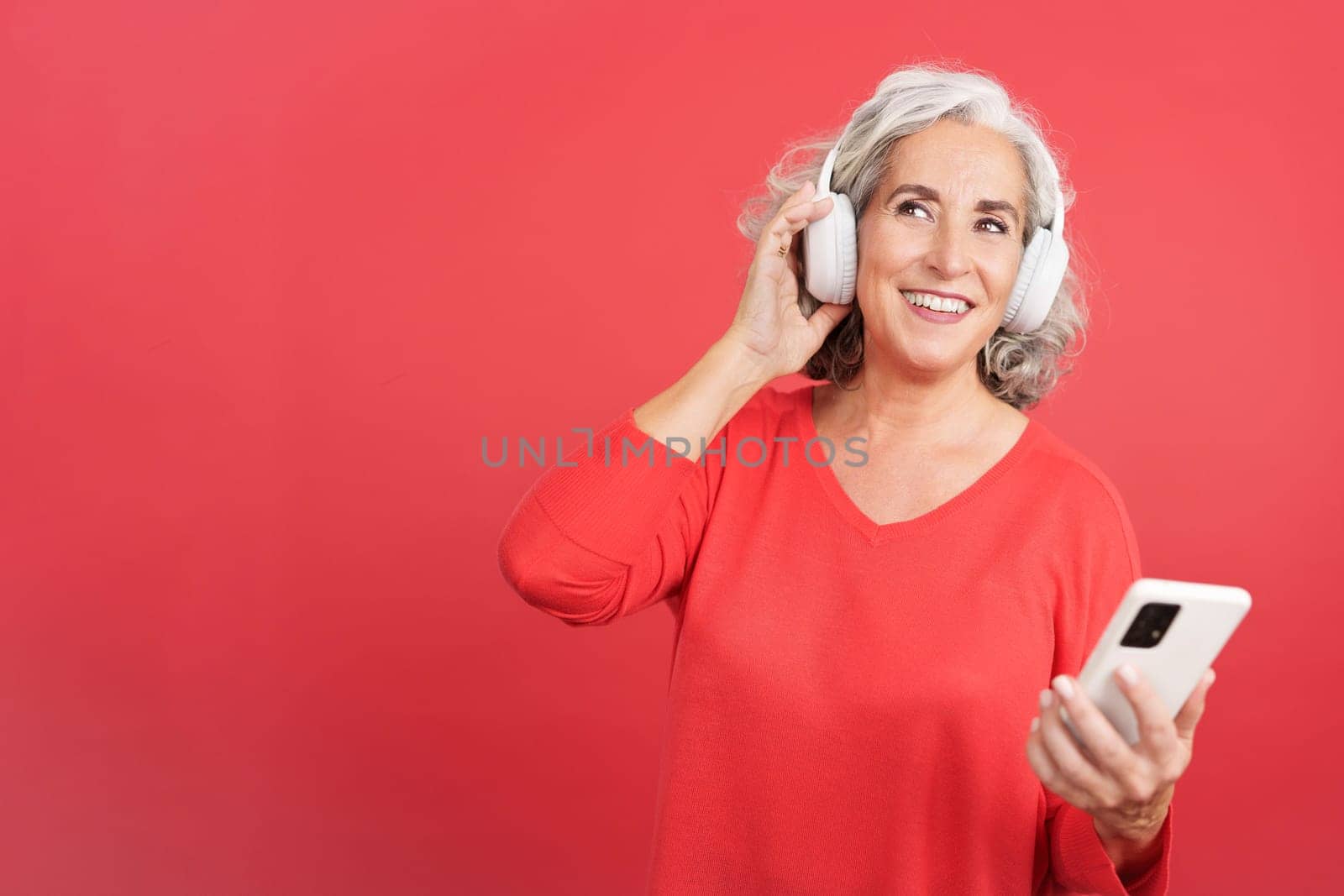 Studio portrait with red background of a distracted woman listening a podcast using the mobile and headphones
