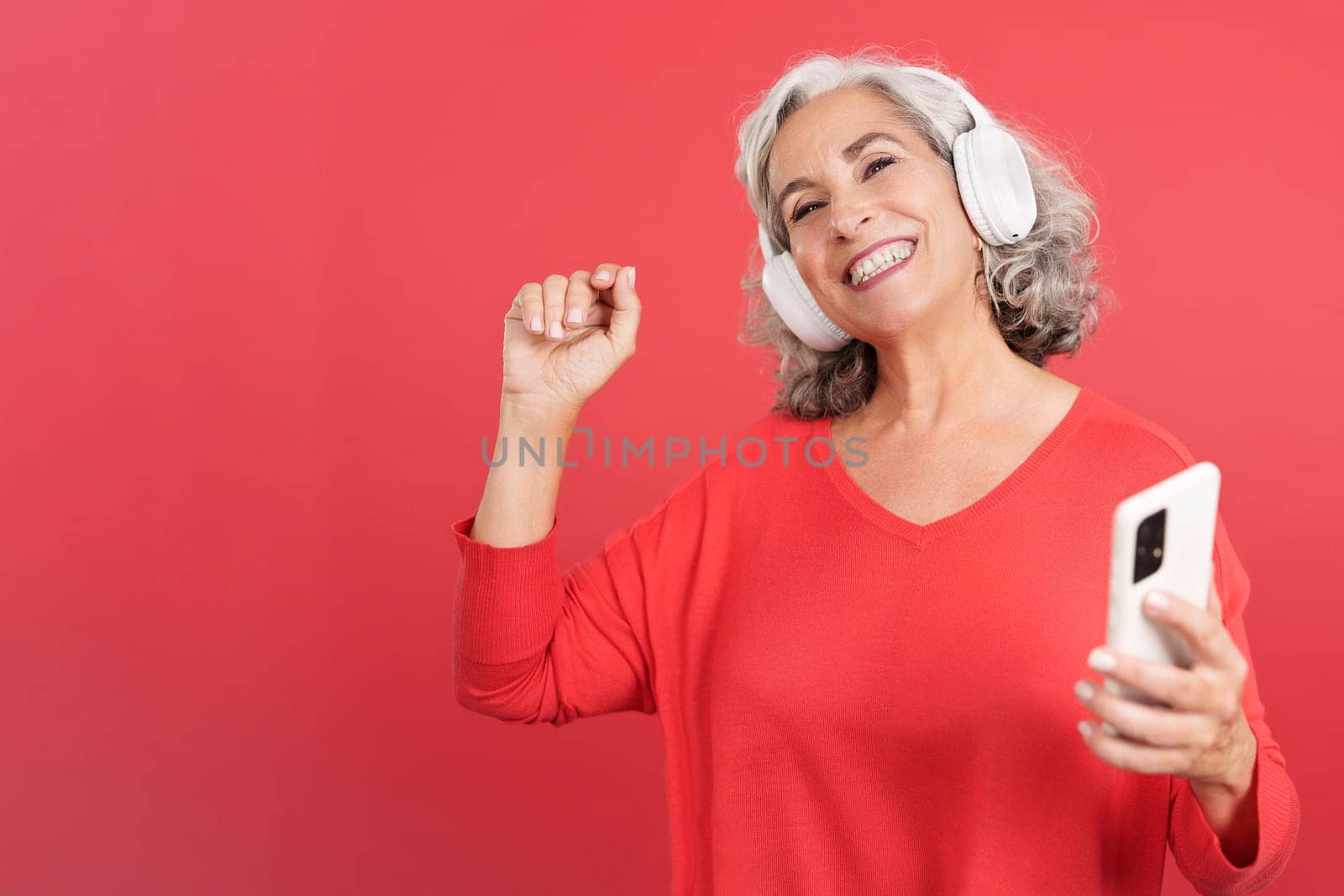 Studio portrait with red background of a happy mature woman dancing and listening to music with a mobile