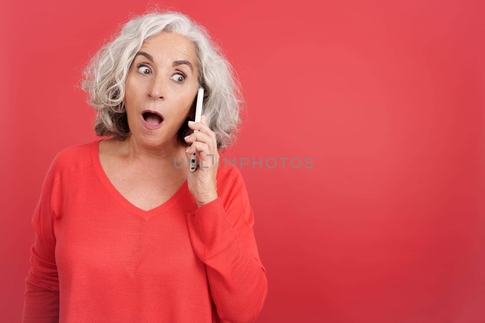 Studio portrait with red background of a surprised mature woman talking to the mobile