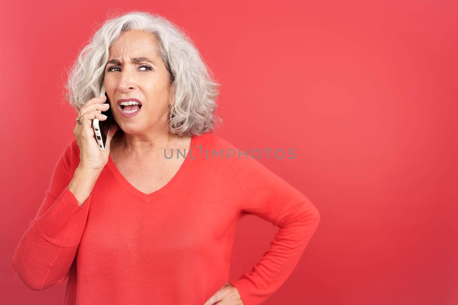 Studio portrait with red background of a mature woman trying to talk on her mobile phone but she can't hear anything