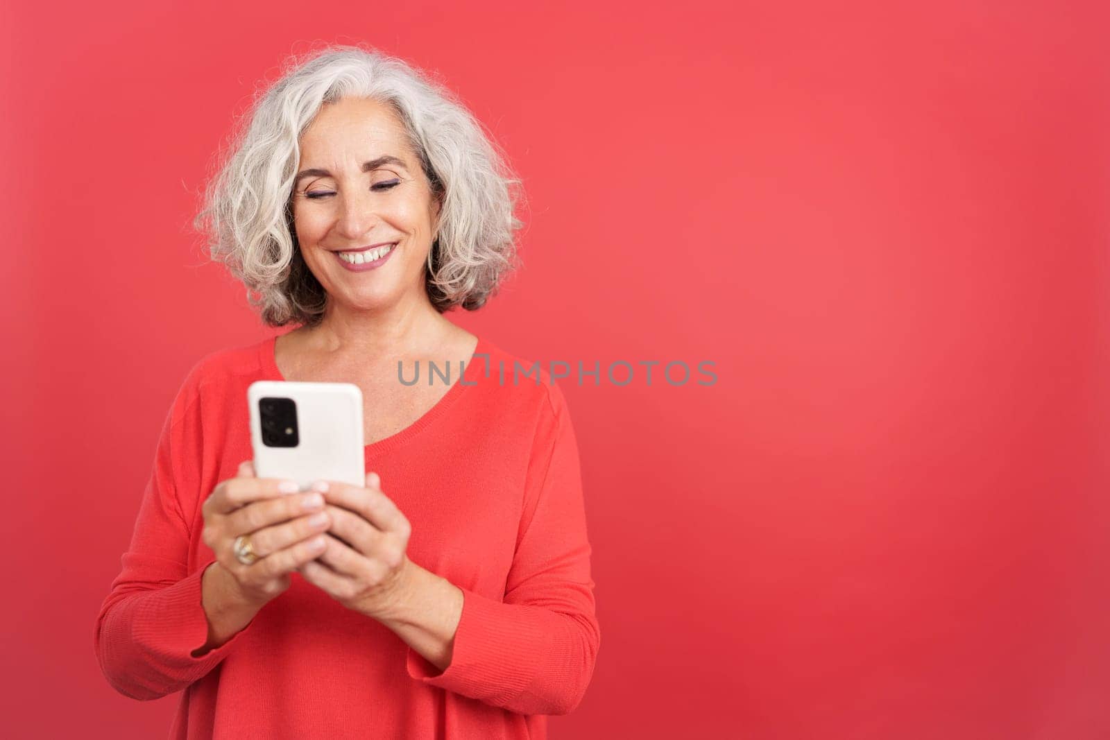 Studio portrait with red background of a mature woman smiling and using the mobile