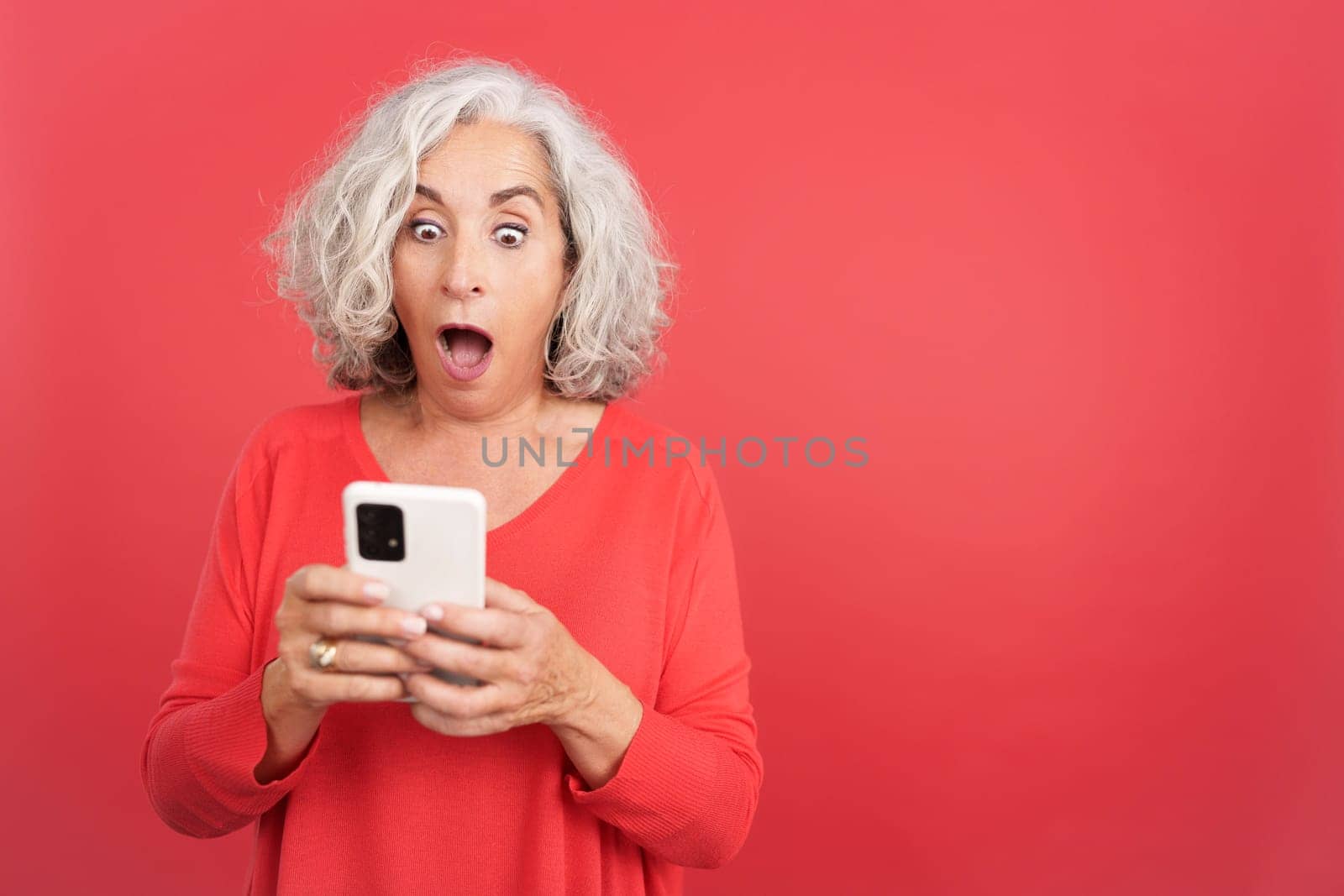 Studio portrait with red background of a surprised mature woman using the mobile phone