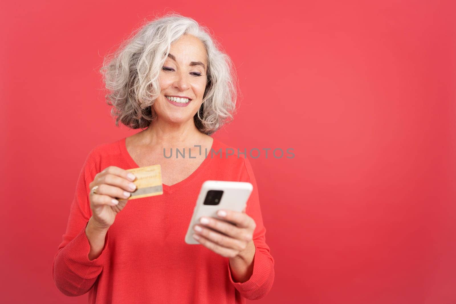 Studio portrait with red background of a mature woman using a credit card to shopping online with the mobile
