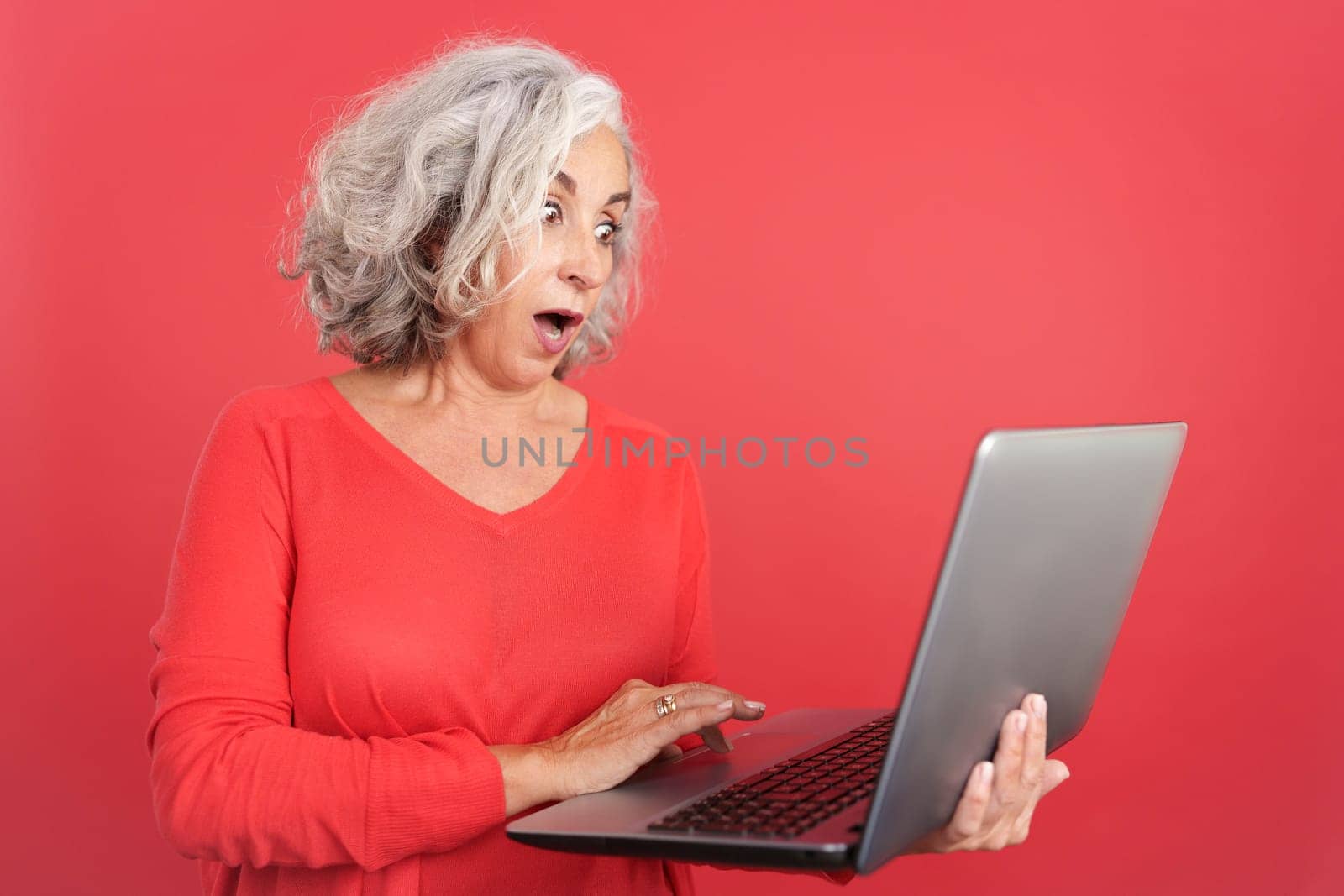 Studio photo with red background of a surprised mature woman standing and using a laptop
