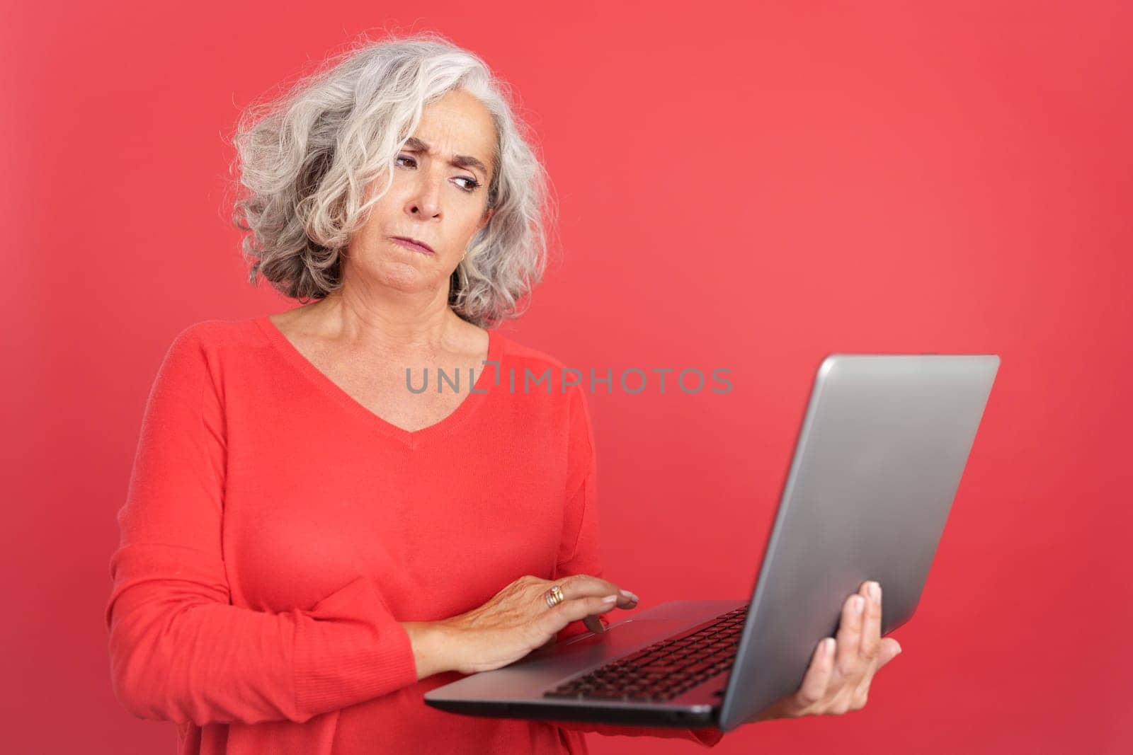 Studio portrait with red background of a worried mature woman standing and using a laptop