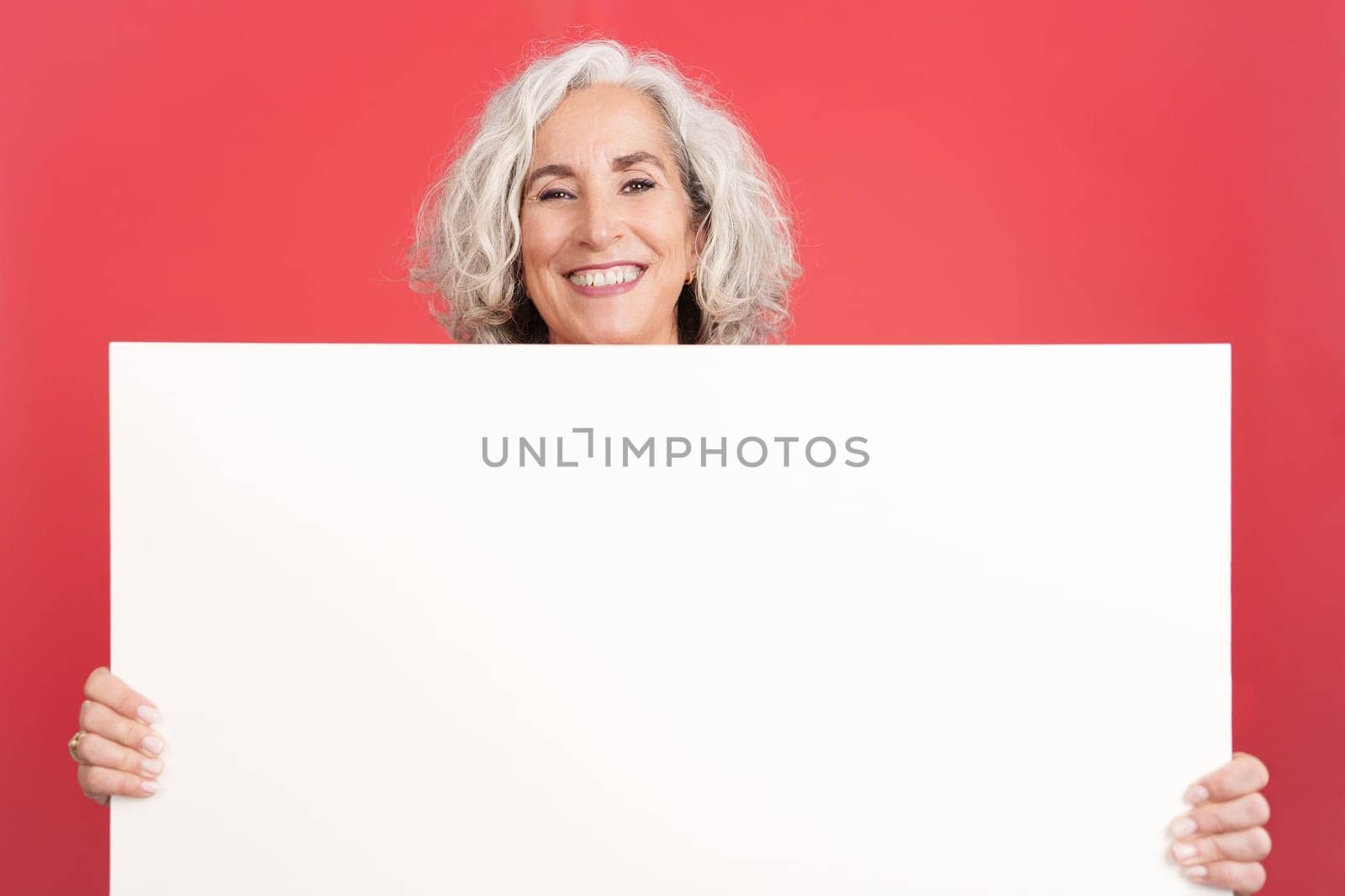 Studio portrait with red background of a mature smiley woman holding a blank panel. Mock up
