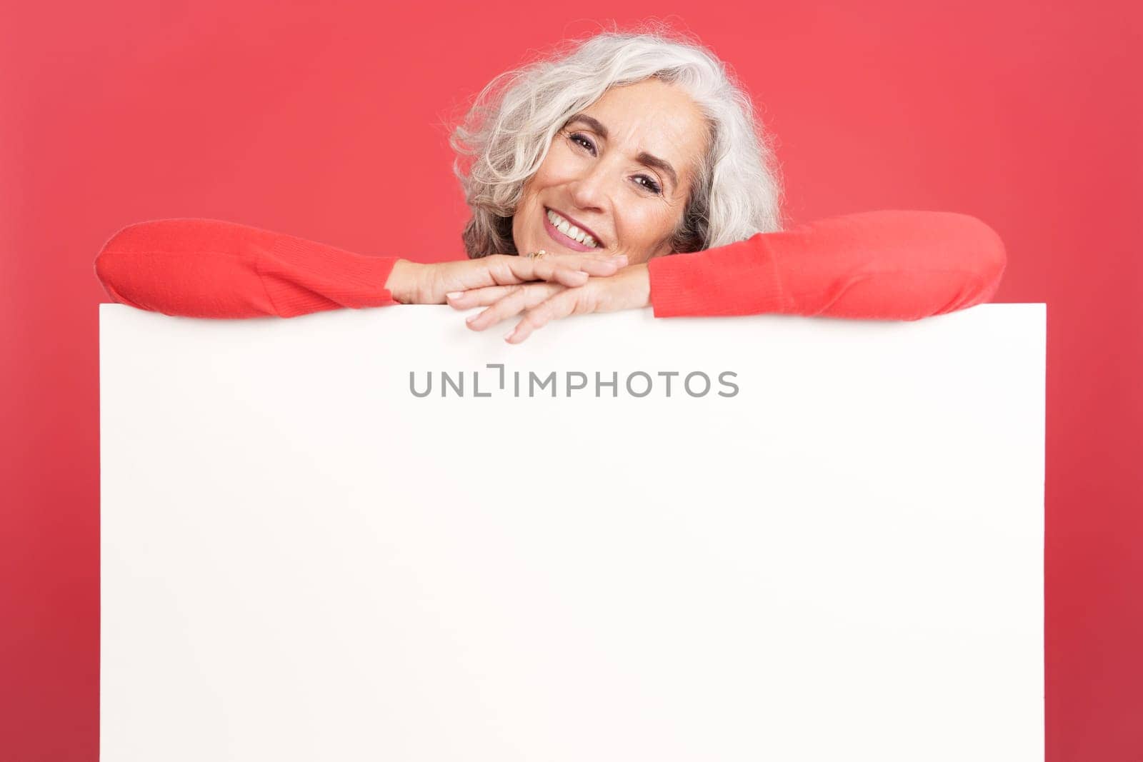 Studio portrait with red background of a relaxed mature woman leaning on a blank panel