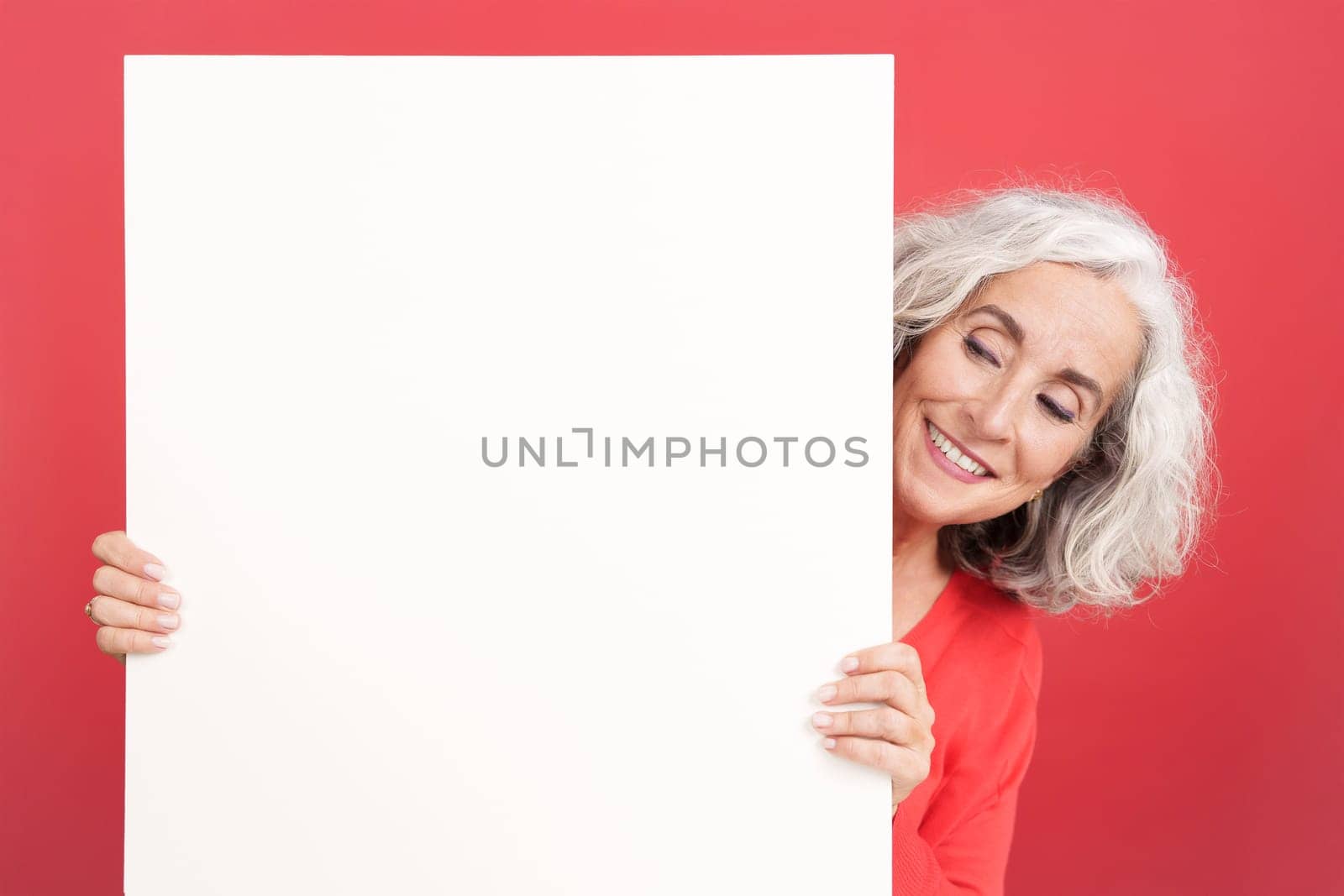 Studio portrait with red background of a mature woman looking at the white board that she is holding