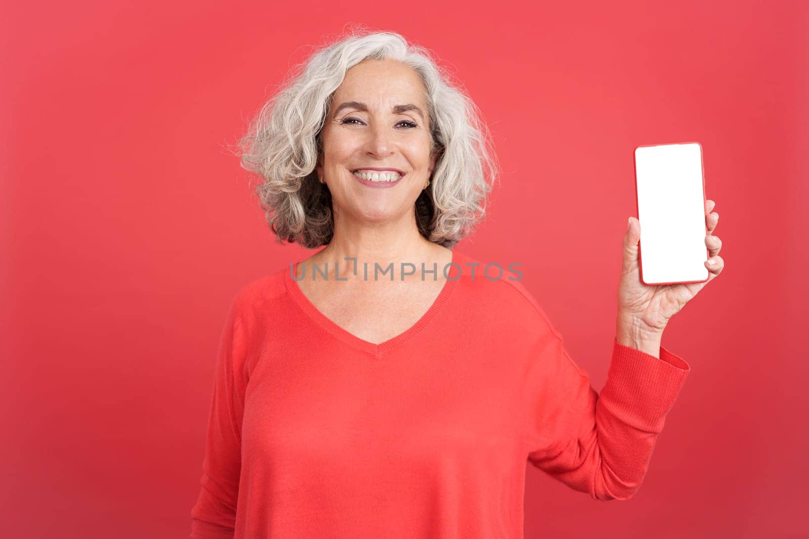 Studio portrait with red background of a mature woman showing the white screen of the mobile, Mock up