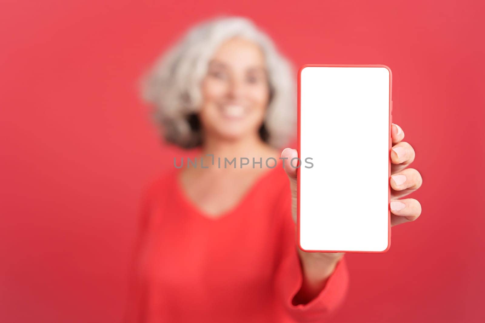 Studio portrait with red background of a blank screen of the mobile held by a mature smiley woman