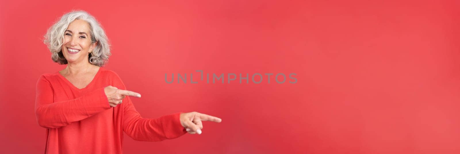 Panoramic Studio portrait with red background of a smiley mature woman pointing to the side