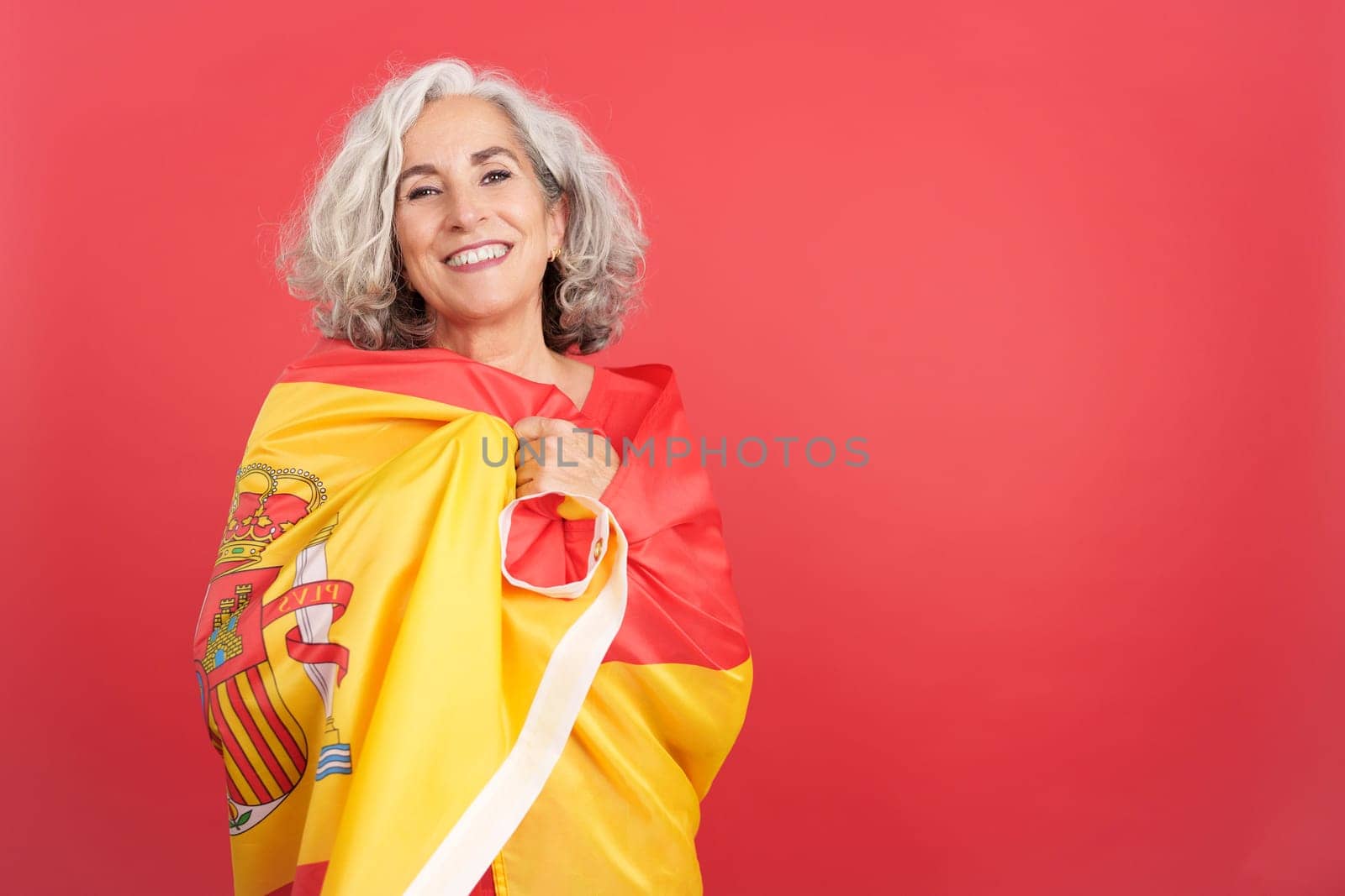 Studio portrait with red background of a smiley mature woman looking at camera while wrapping with a spanish national flag