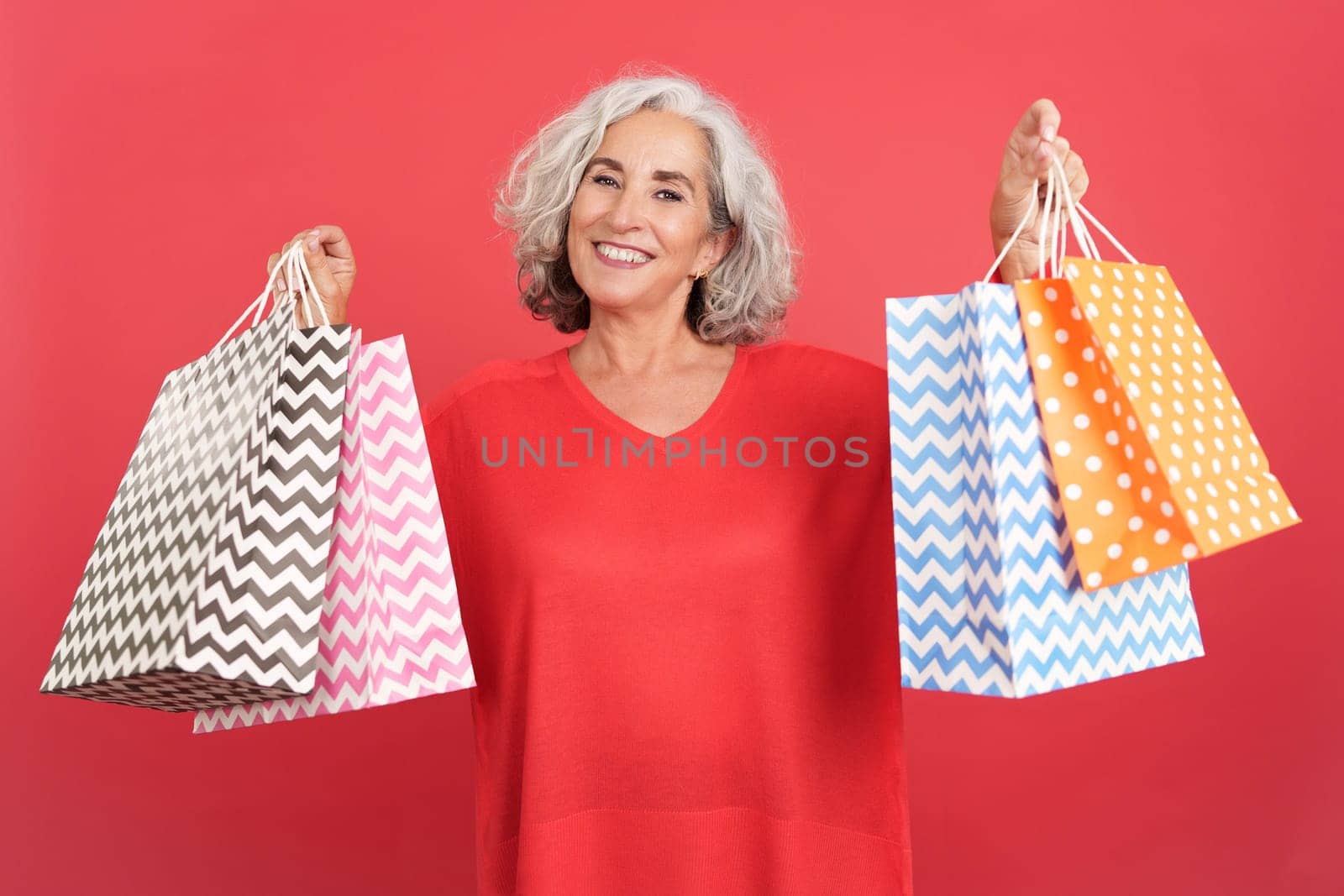 Studio portrait with red background of a happy mature woman standing showing shopping bags