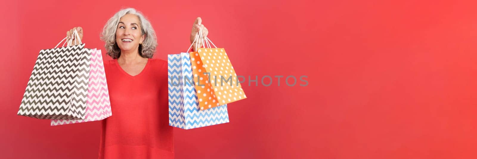 Studio portrait with red background of a happy mature woman looking up while holding shopping bags