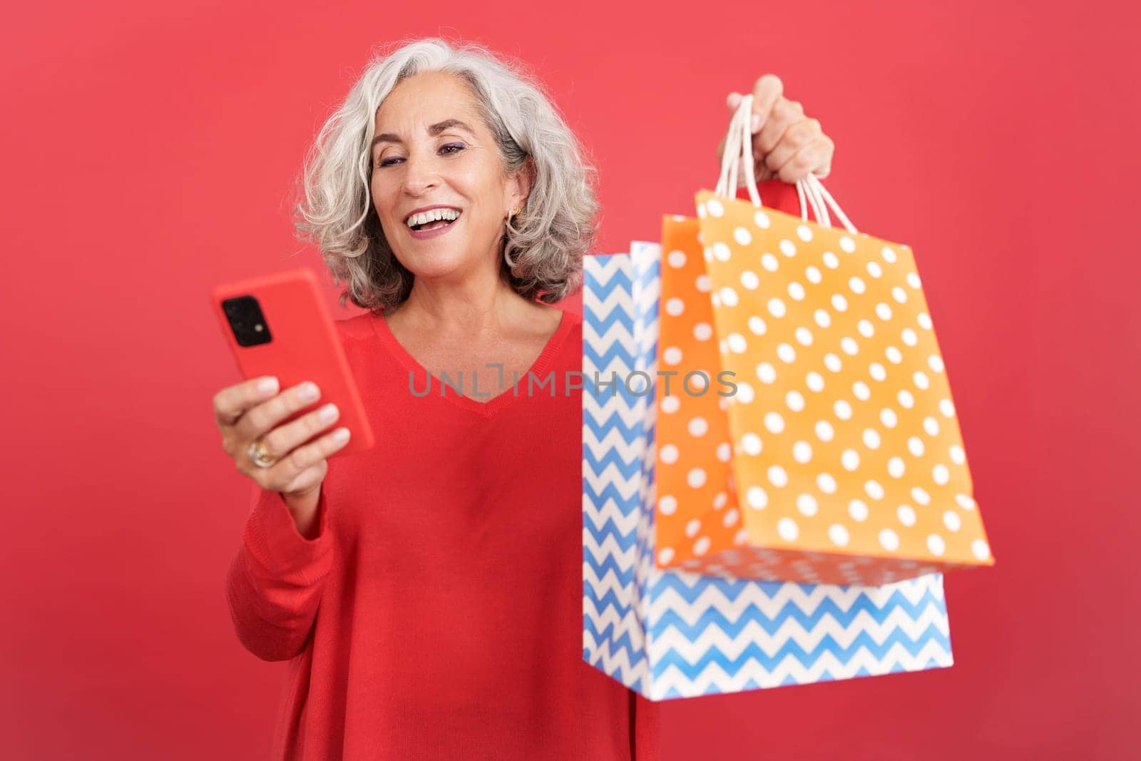 Studio portrait with red background of a mature woman using a mobile while holding shopping bags