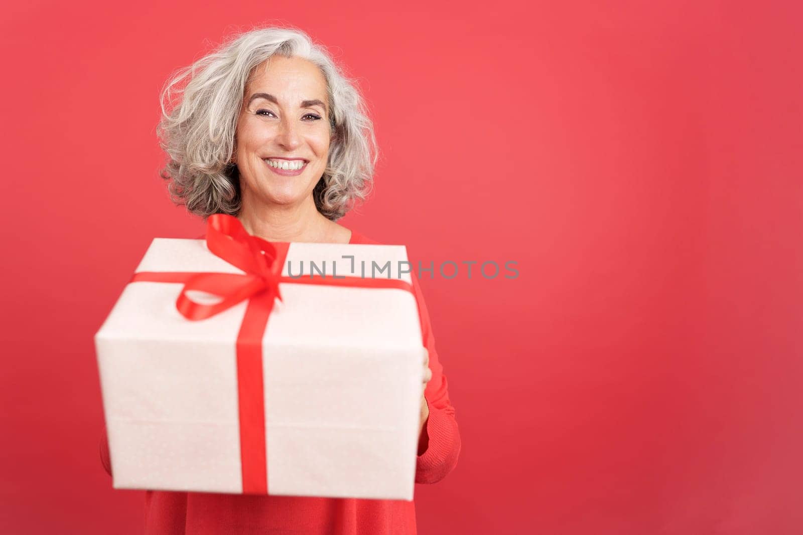 Studio portrait with red background of a happy mature woman giving a gift