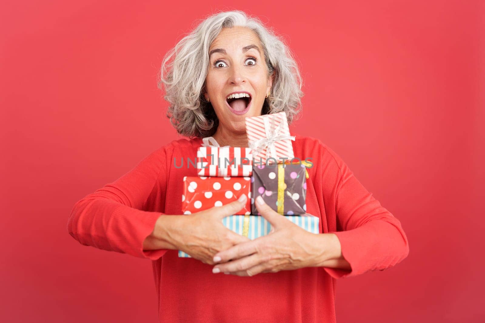 Studio portrait with red background of an excited mature woman receiving many gifts