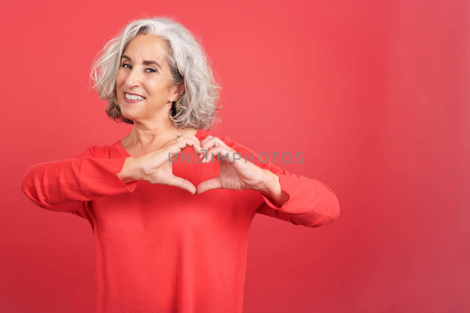 Studio portrait with red background of a mature woman depicting a heart in the shape of her fingers