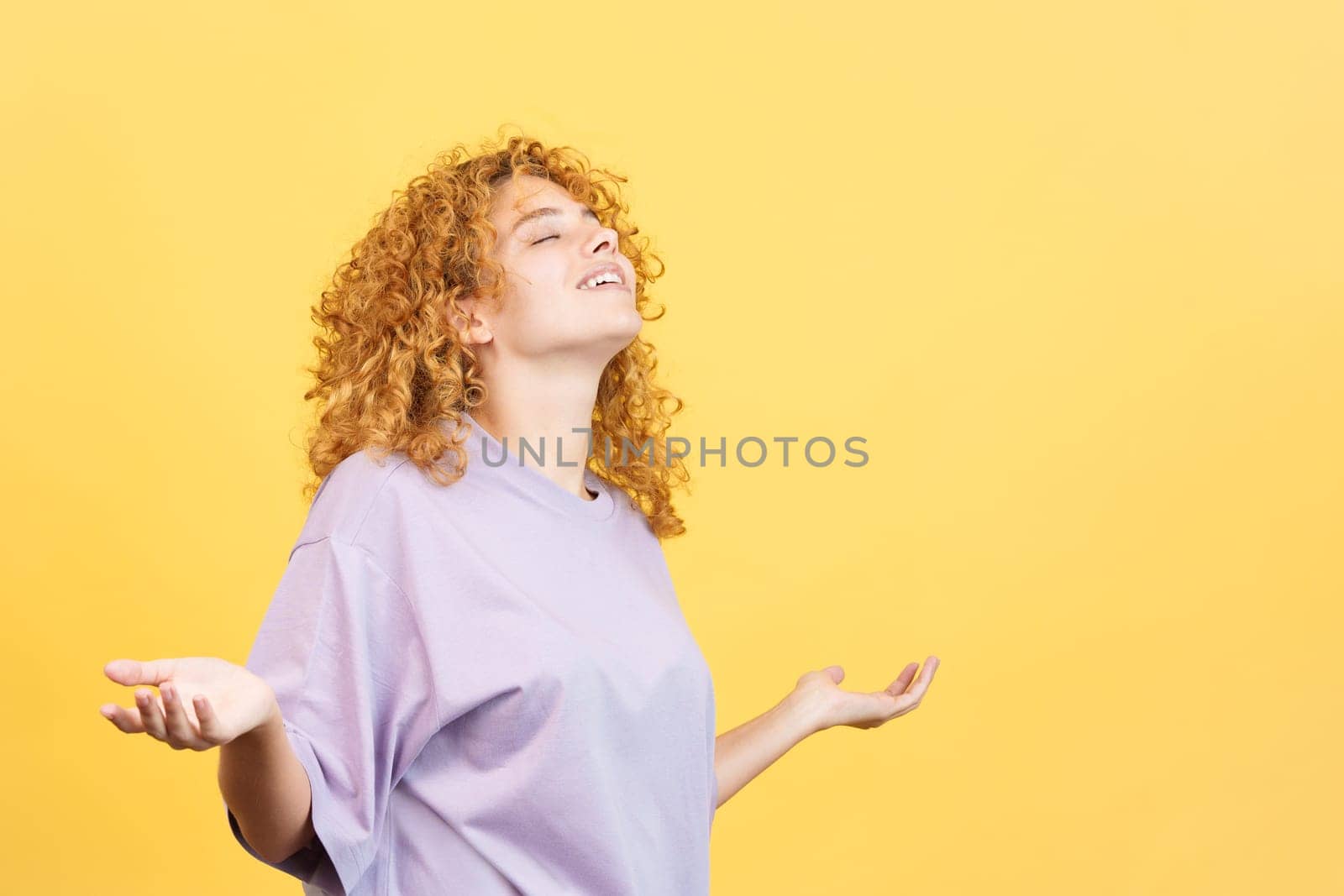 Studio image with yellow background of a woman with curly hair standing with closed eyes and open arms gesturing freedom
