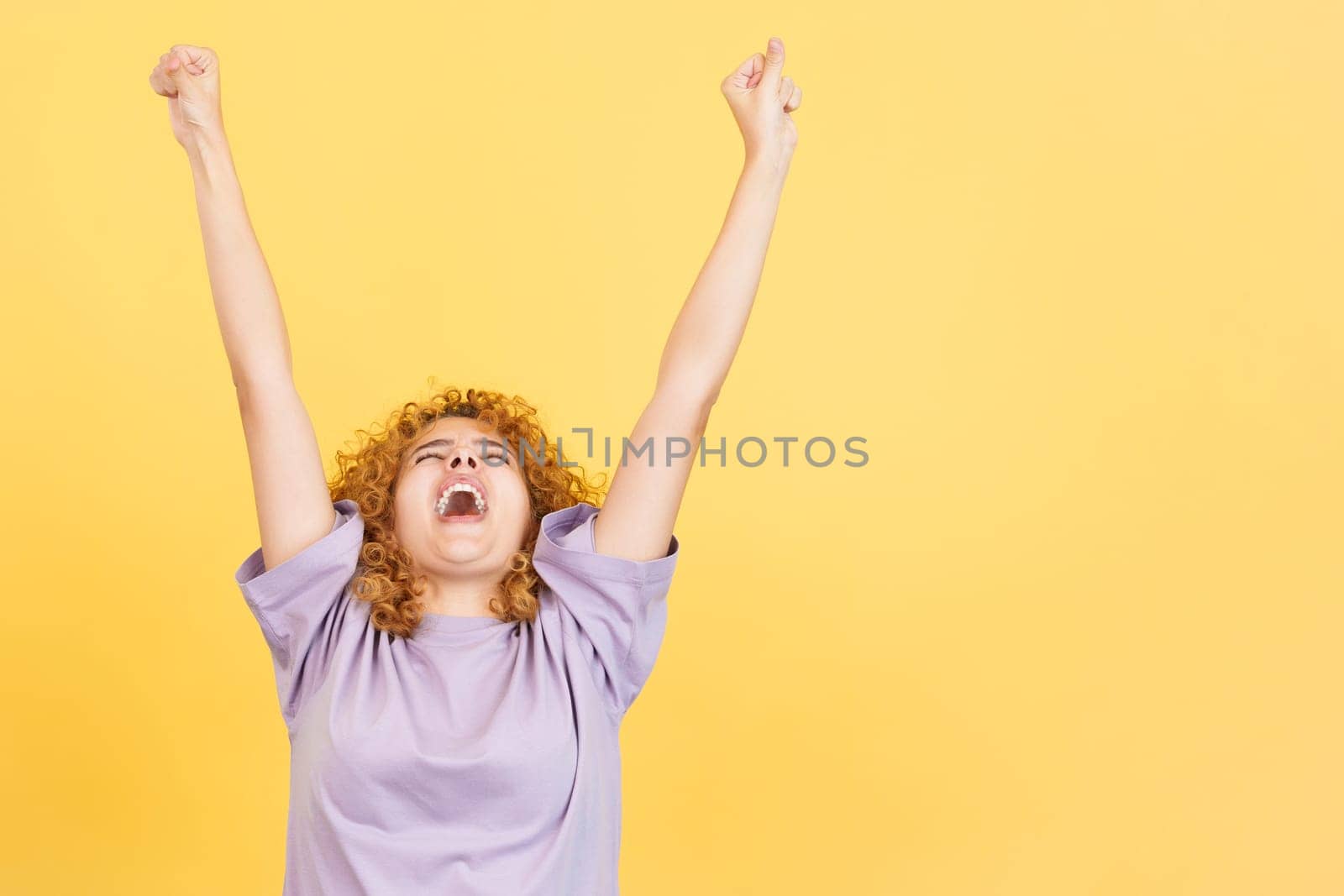 Studio image with yellow background of a happy woman with curly hair celebrating while raising the arms and shouting