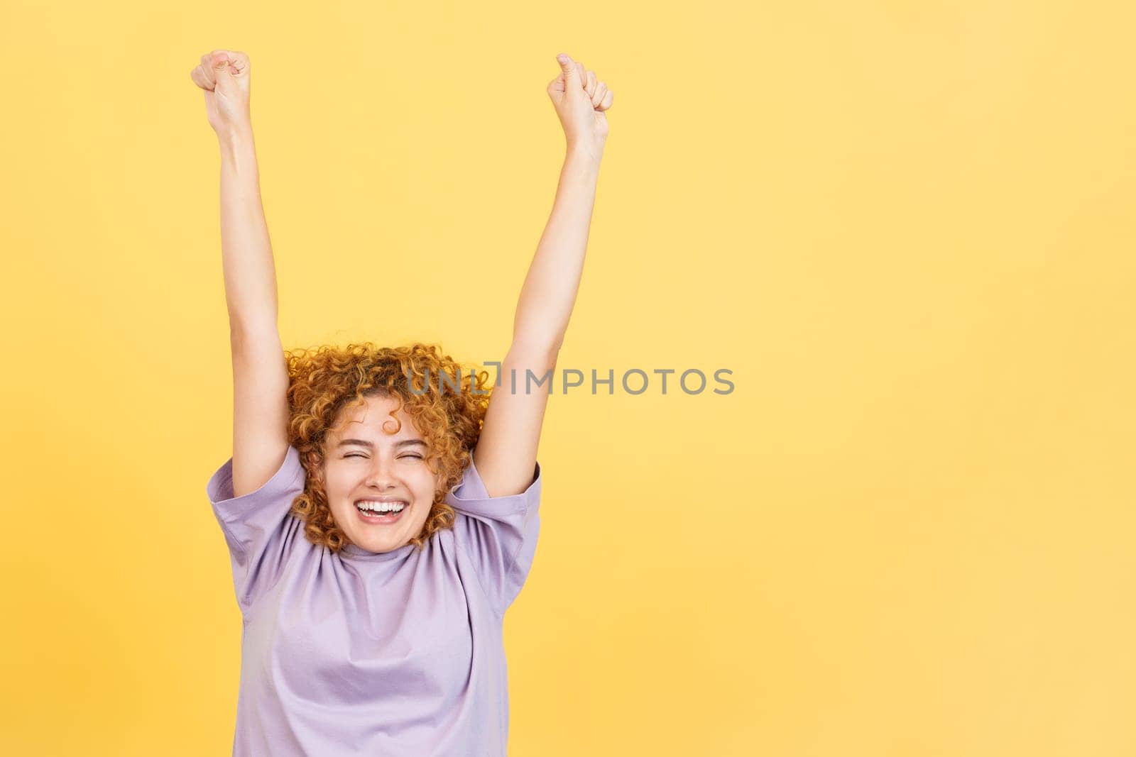 Studio image with yellow background of a happy woman with curly hair celebrating while raising the arms and smiling