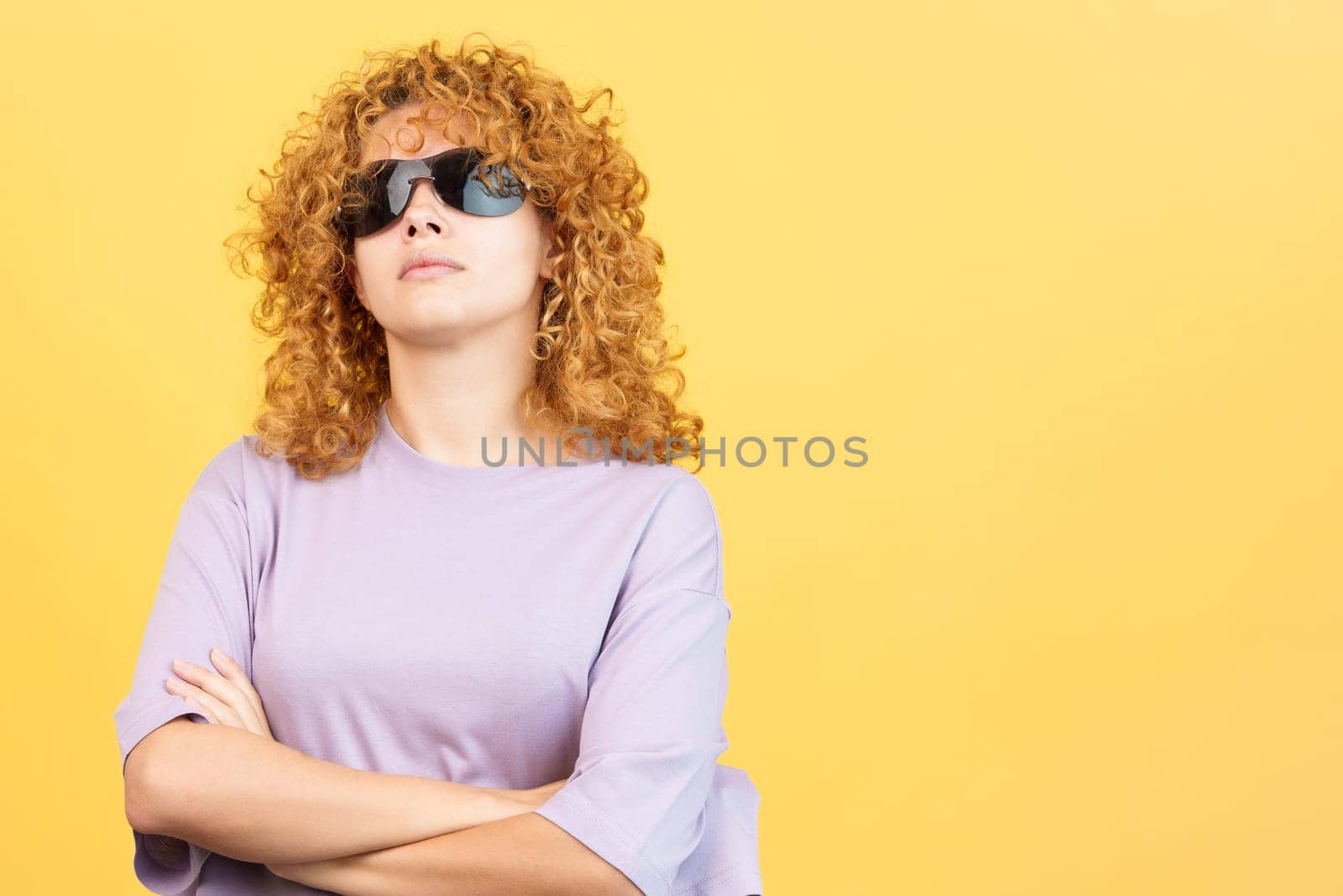Studio image with yellow background of a rude woman with curly hair and sunglasses standing with arms crossed