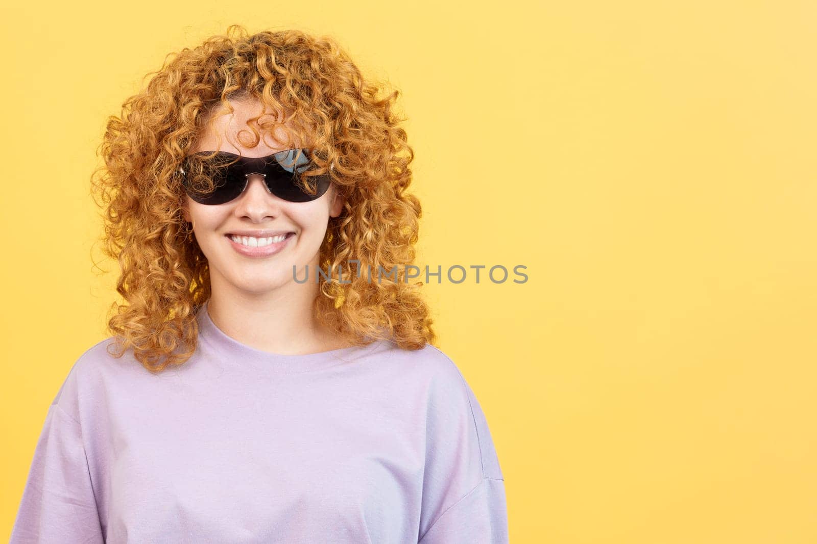 Studio image with yellow background of a happy woman with curly hair and sunglasses looking at the camera