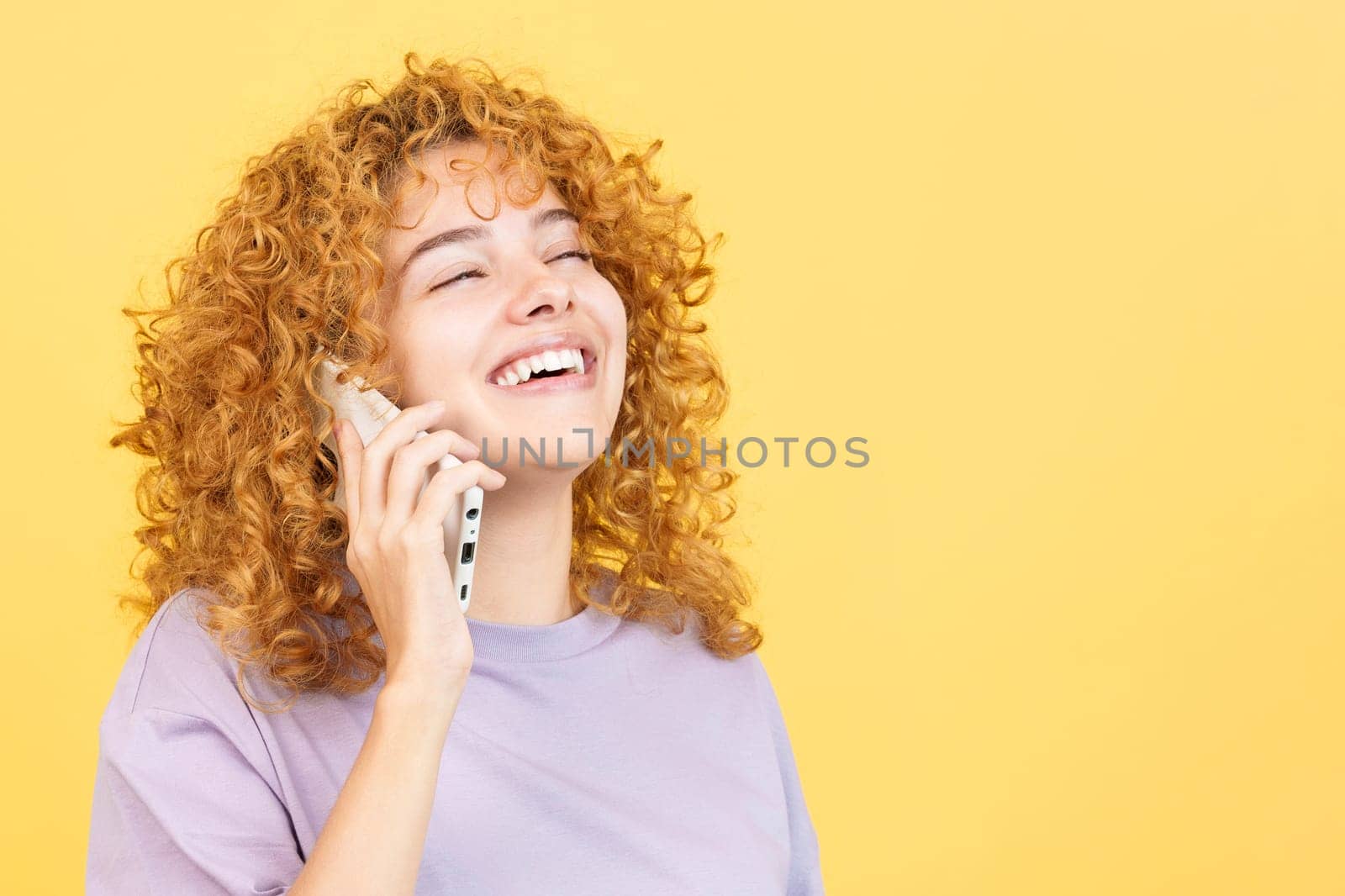 Studio image with yellow background of a beauty woman with curly hair talking to the mobile while smiling with eyes closed