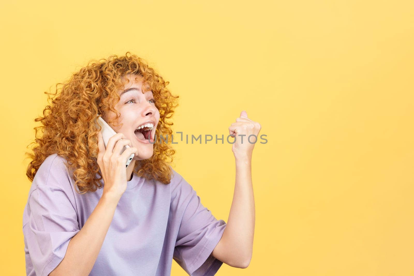 Studio image with yellow background of an excited woman with curly hair celebrating and screaming while taking to the mobile