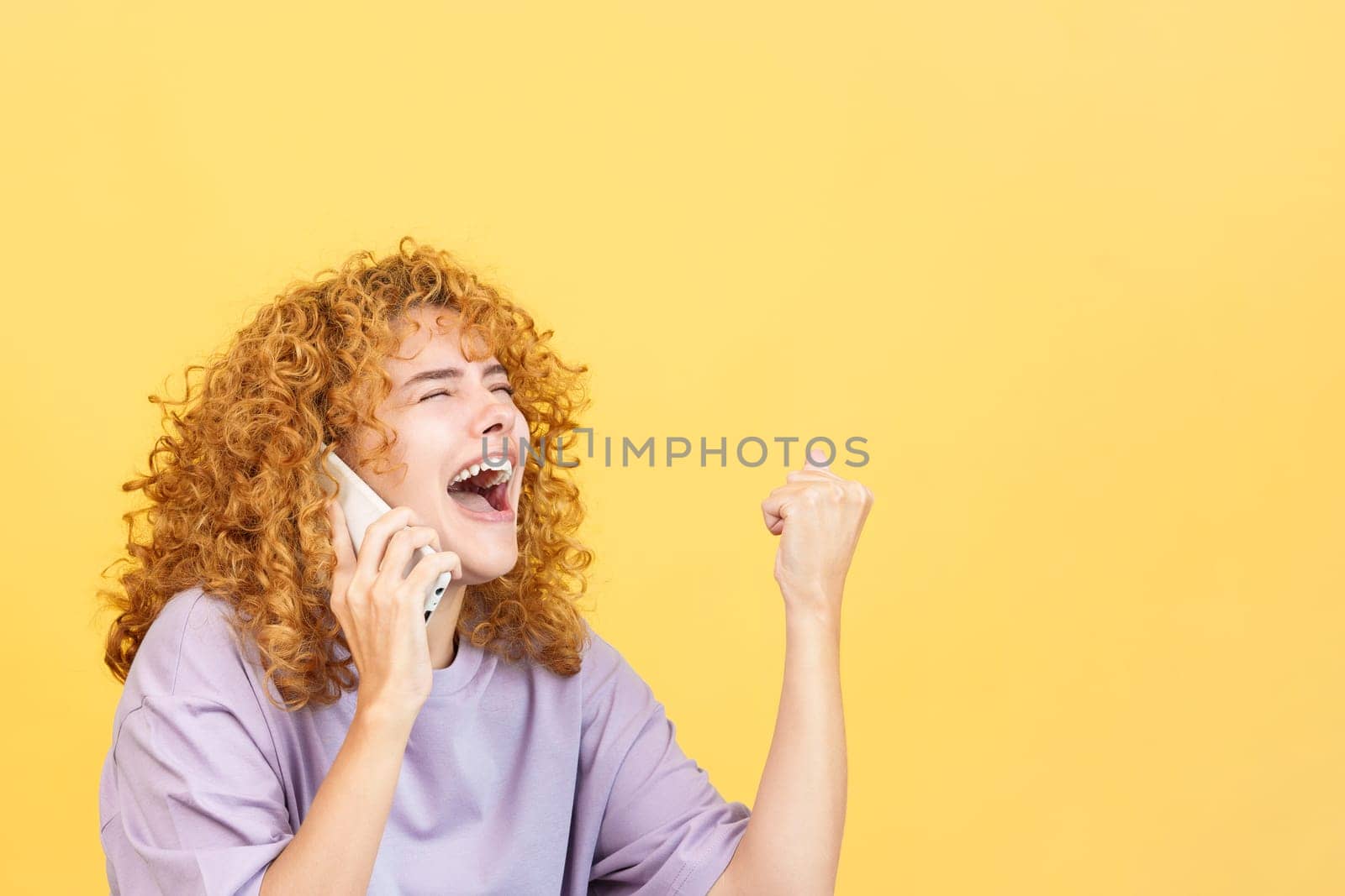 Studio image with yellow background of a excited and happy woman with curly hair celebrating while taking to the mobile