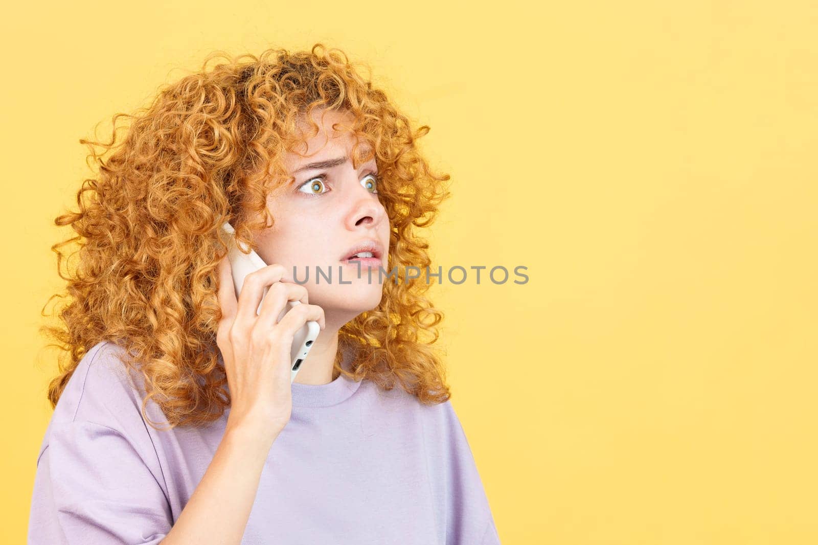 Studio image with yellow background of a frightened woman with curly hair talk to the mobile phone