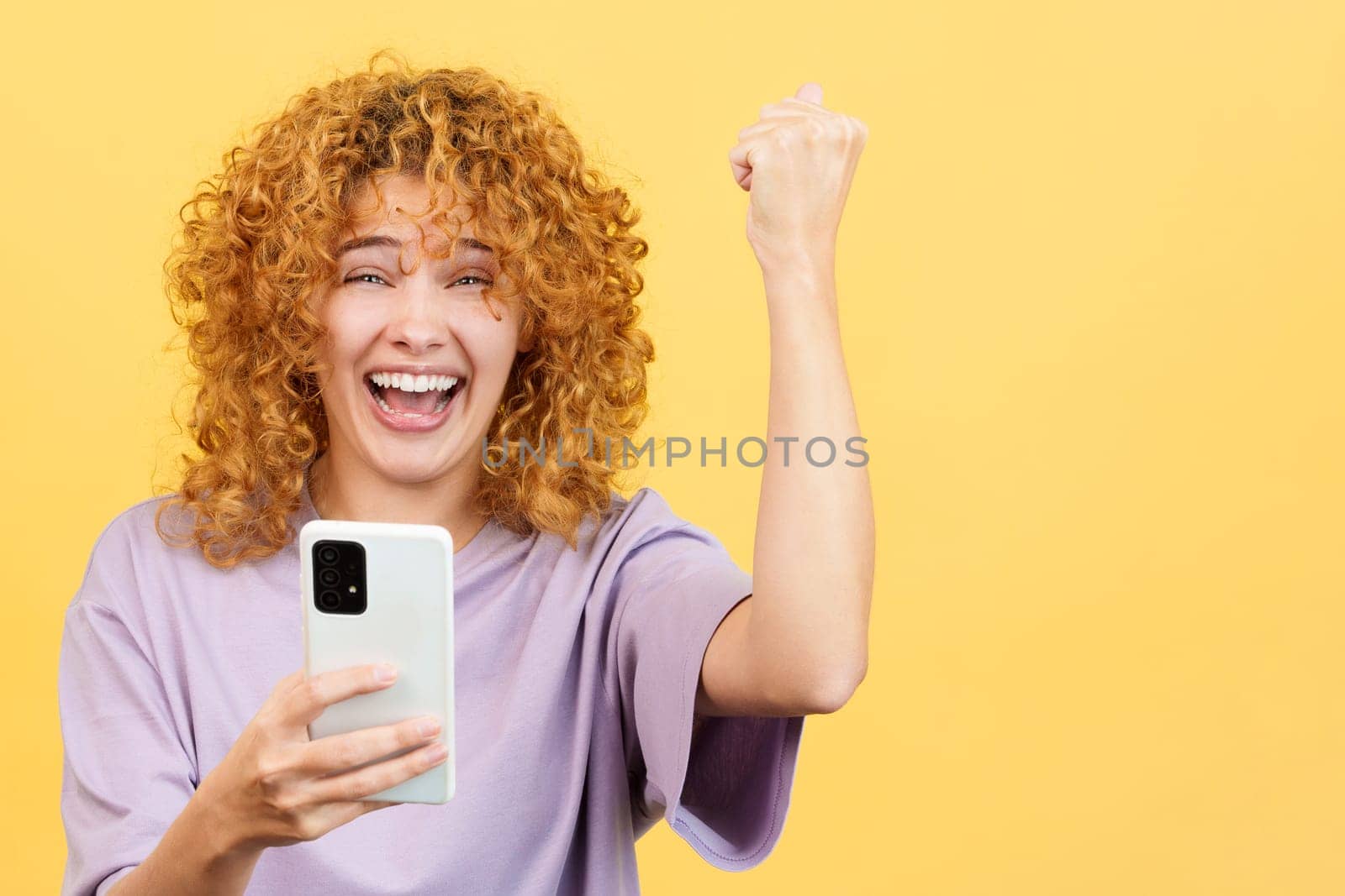 Studio image with yellow background of an excited woman with curly hair celebrating while using the mobile phone