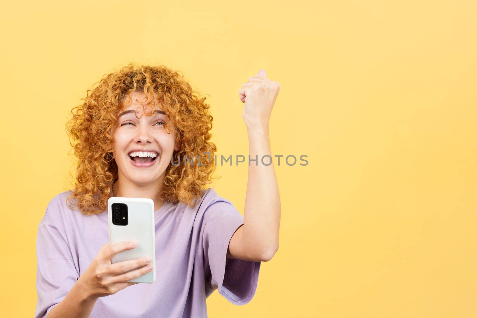 Studio image with yellow background of a happy woman with curly hair celebrating while using the mobile phone