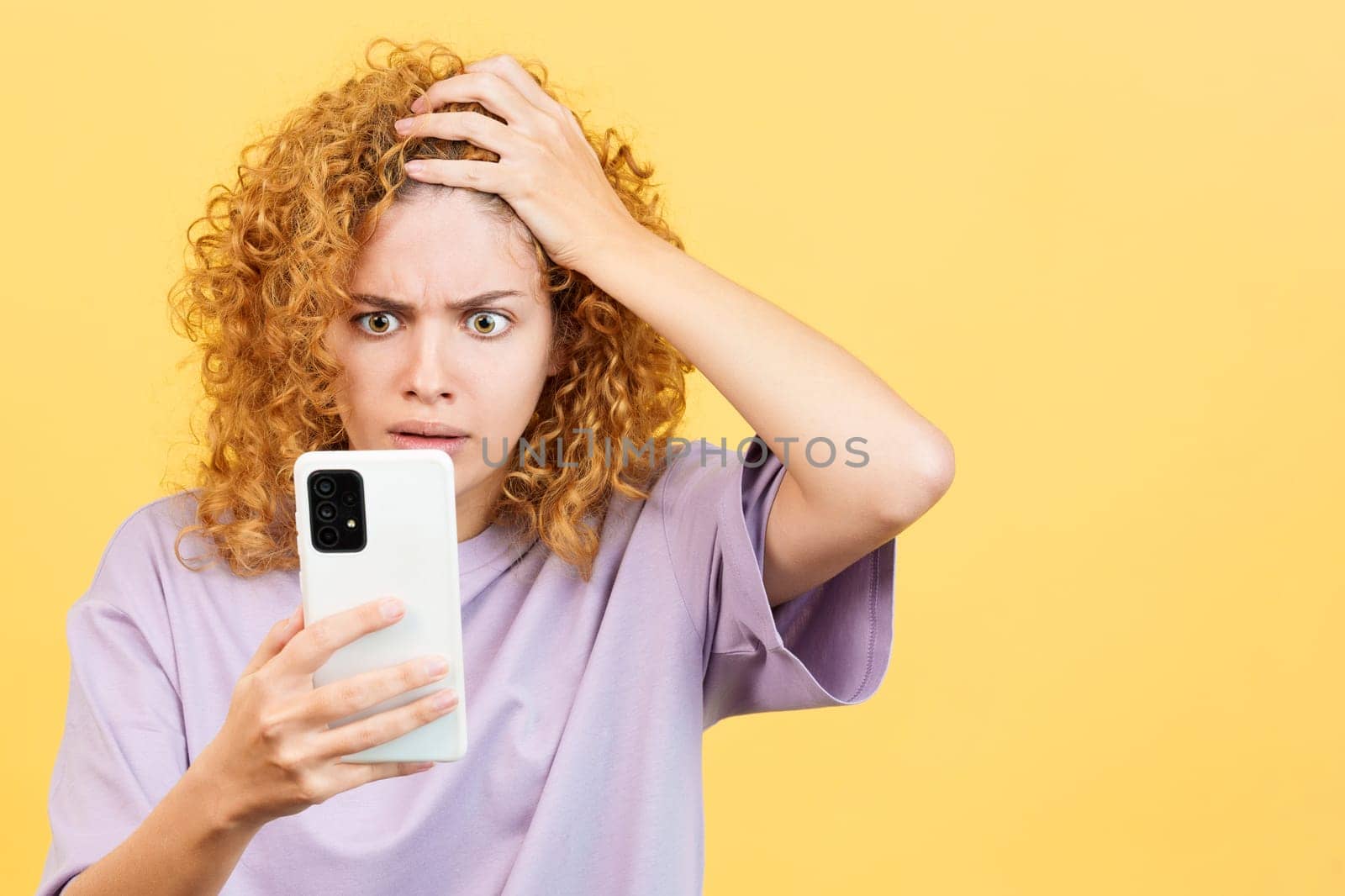 Studio image with yellow background of a desperate woman with curly hair looking at mobile phone and gesturing