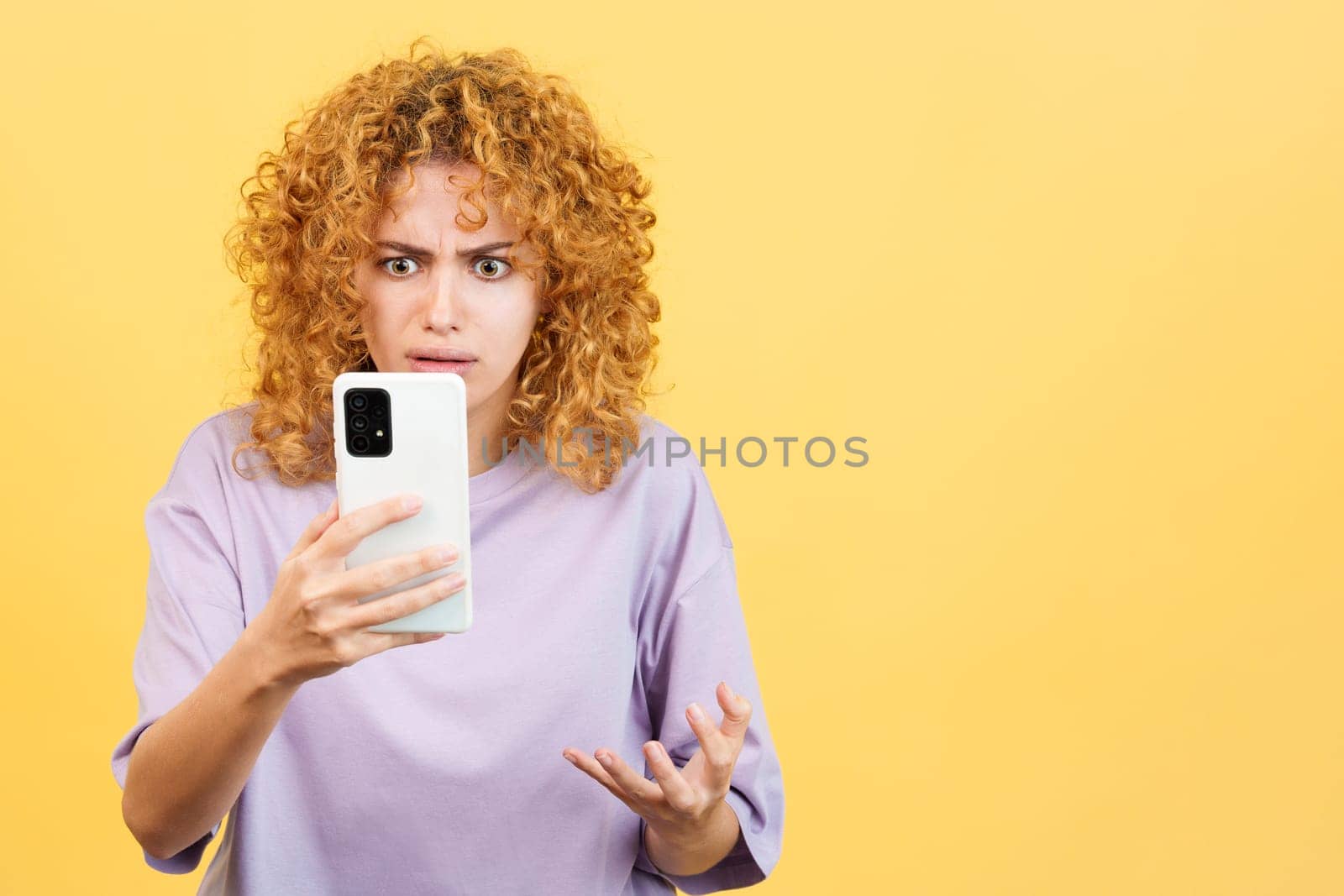 Studio image with yellow background of a upset and worried woman with curly hair using a mobile