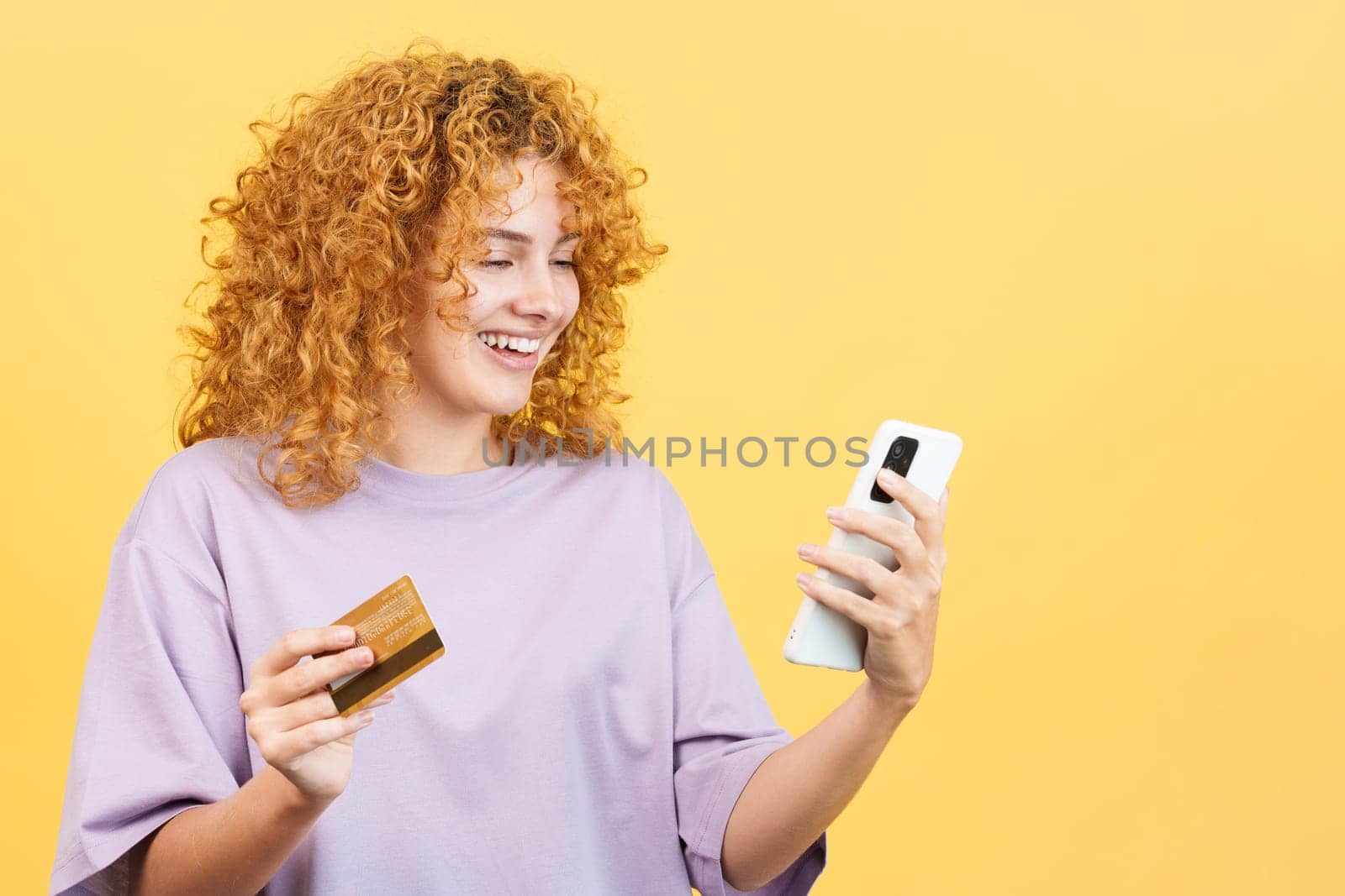 Studio image with yellow background of a smiley woman with curly hair using a card to shopping online with the mobile